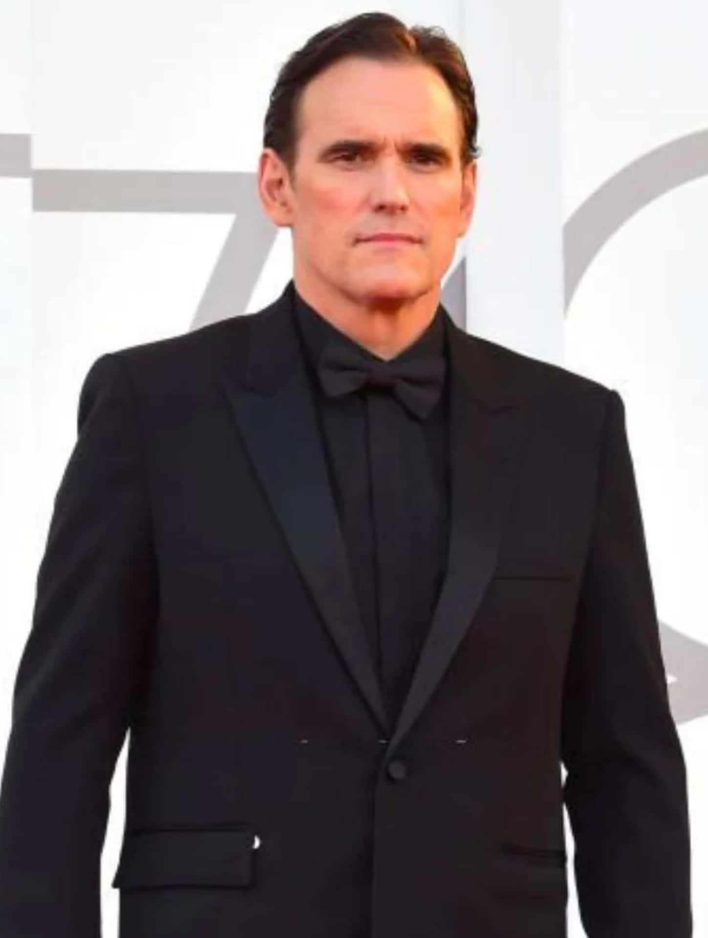matt dillon 桌布 壁紙 — 一位身穿俐落黑色燕尾服搭配領結的男士，在帶有幾何灰色點綴的極簡白色背景前從容佇立。
