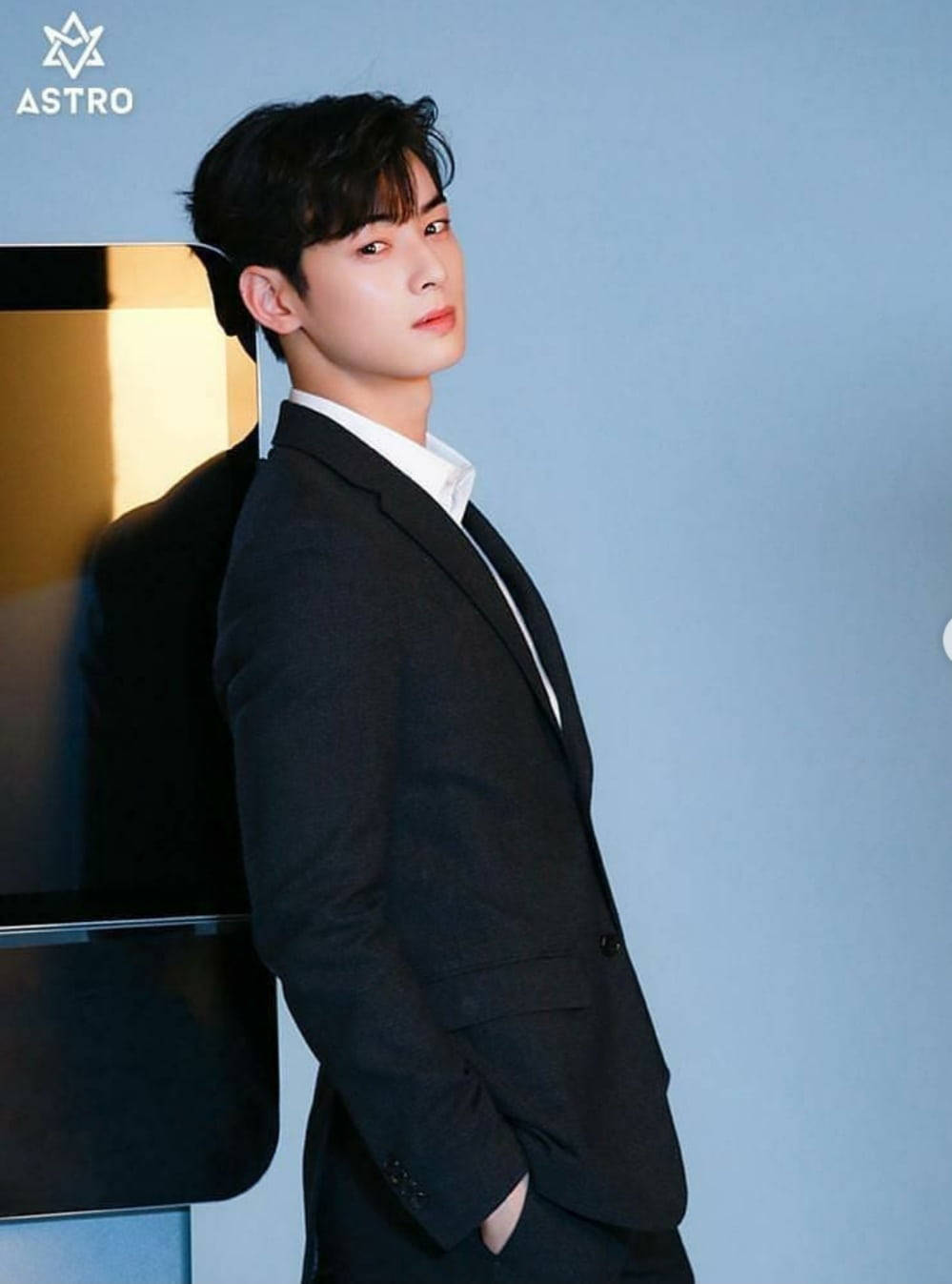 Eunwoo 桌布 壁紙 — 一位身穿俐落黑色西裝的年輕男子，靠著現代金色和黑色結構，背景是柔和的藍色牆壁。