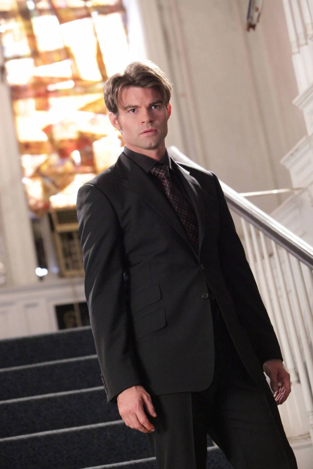 Daniel Gillies 桌布 壁紙 — 一位身穿俐落黑色西裝的男子自信地站在華麗的樓梯上，樓梯有精緻的欄杆和溫暖的金色燈光。