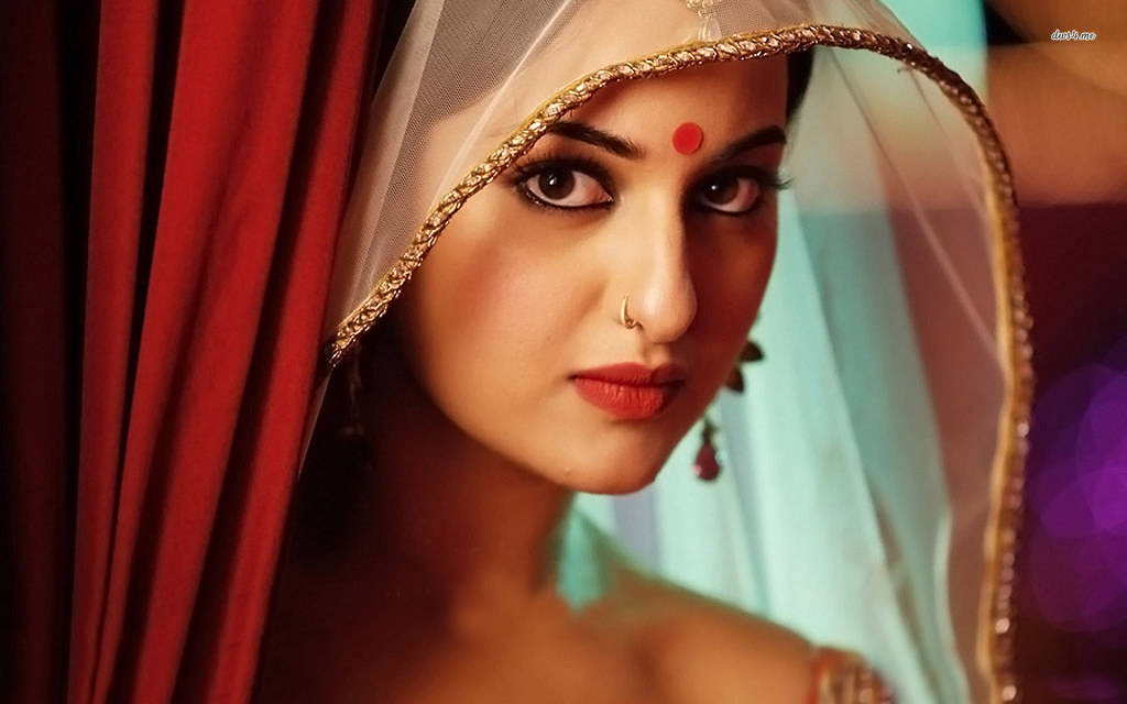 Sonakshi Sinha 桌布 壁紙 — 一位身穿傳統印度新娘服飾、額頭點有紅色吉祥痣、戴著金線刺繡頭紗的女子，從華麗的紅色窗簾後探出頭來。
