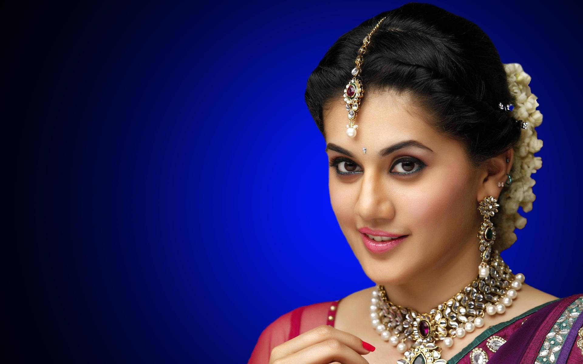 Taapsee Pannu 桌布 壁紙 — 一位身穿傳統印度服飾、佩戴精緻珠寶的女性，在深藍色漸變背景前溫柔微笑。