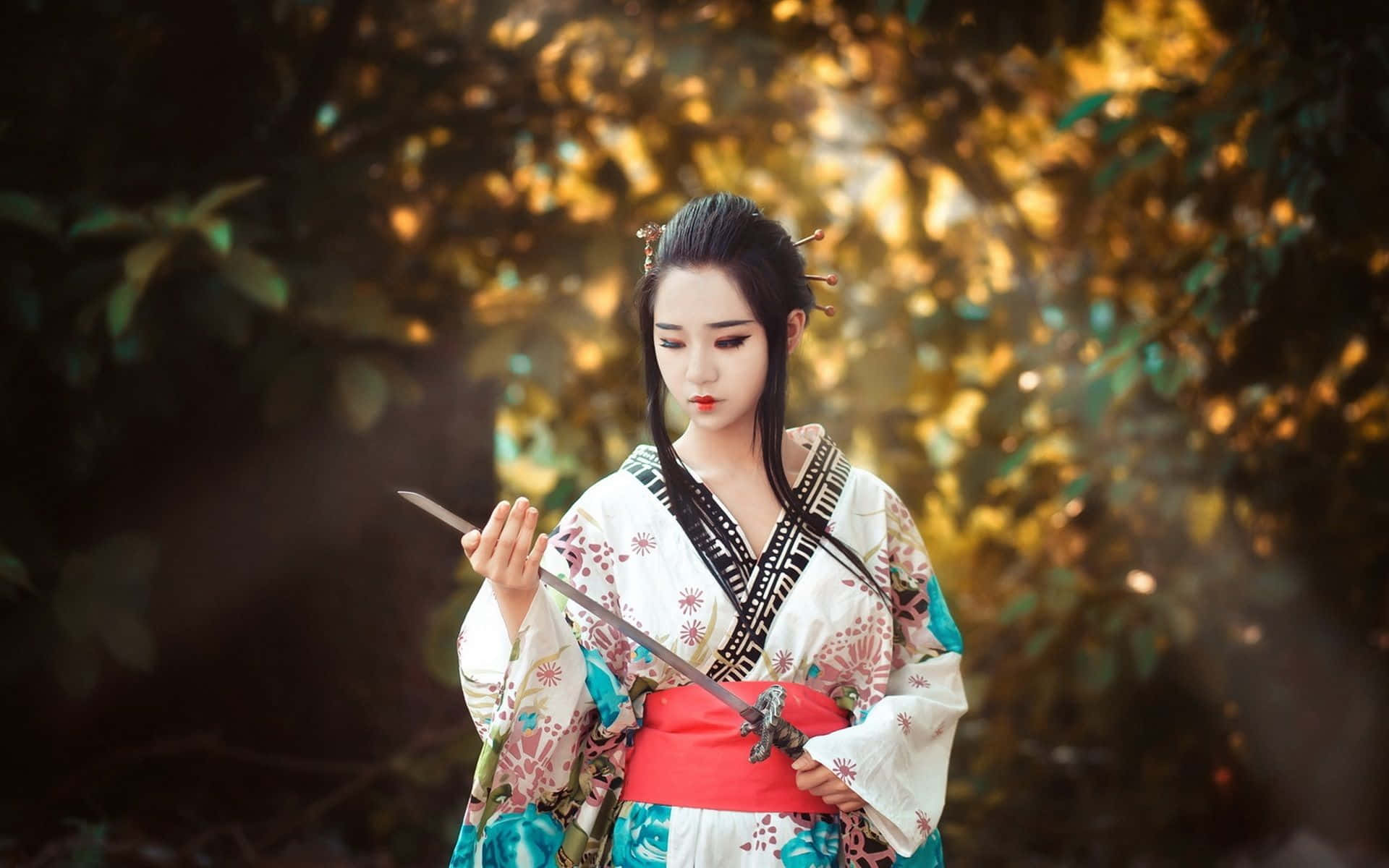 Japanese Women 桌布 壁紙 — 一位身穿傳統日本服飾的女性在陽光明媚、金色樹葉的森林中手持武士刀。