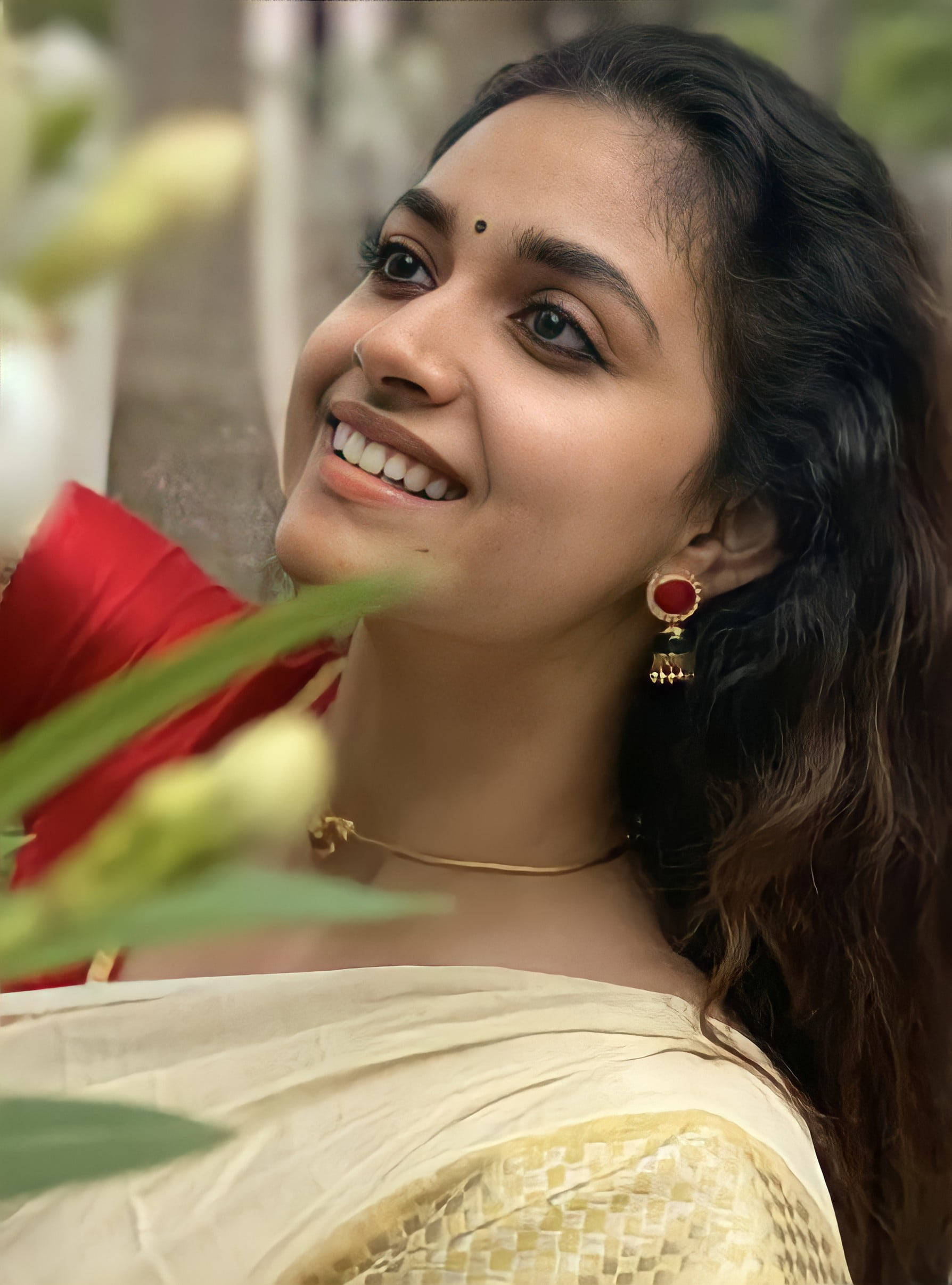telugu 桌布 壁紙 — 一位身穿傳統服飾、光彩照人的女性在柔和模糊的綠色植物和紅色布料中溫暖微笑。