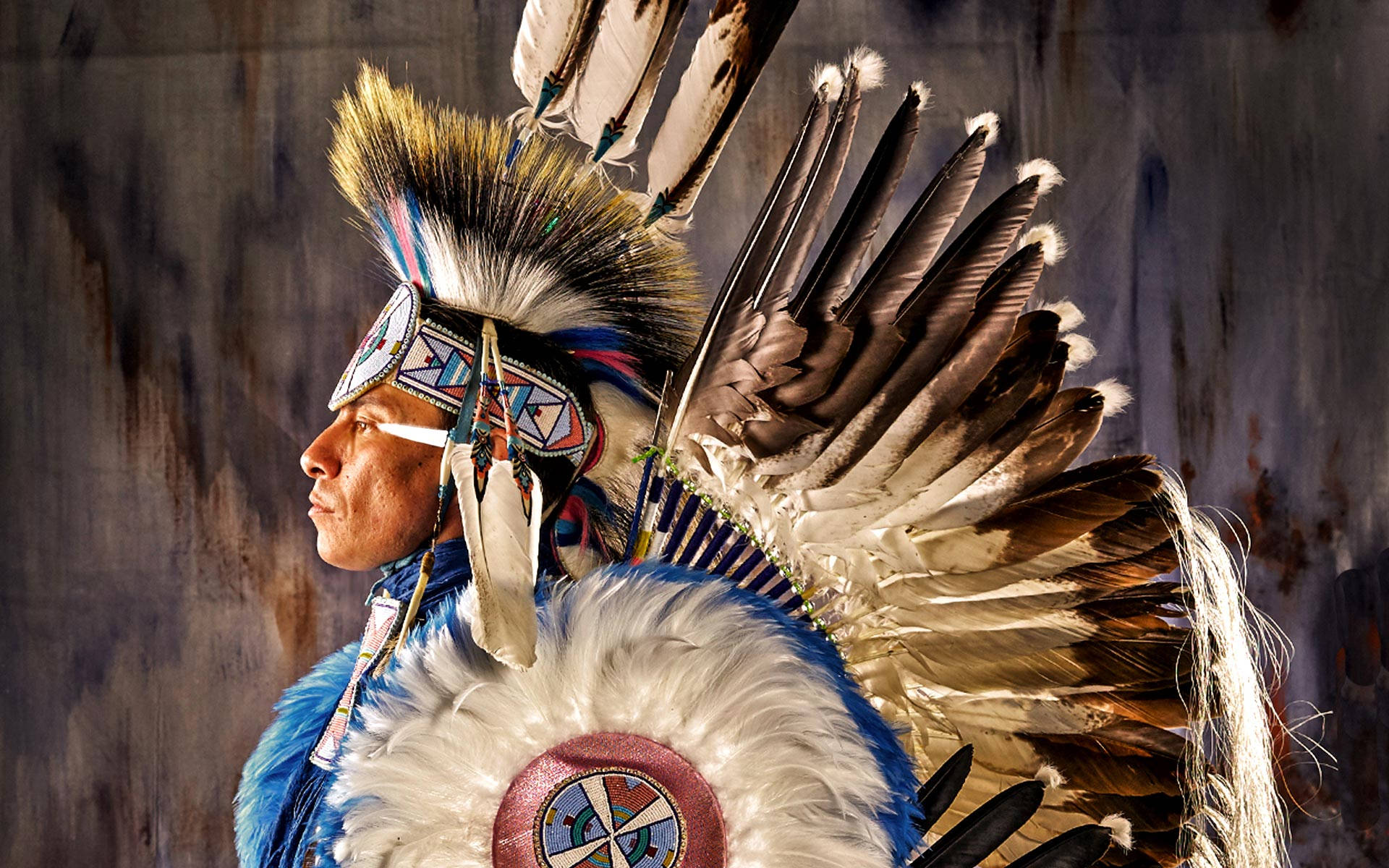 Native American 桌布 壁紙 — 一位身穿傳統服飾、頭戴羽毛頭飾的美洲原住民男子，在風化的木質背景下若有所思地凝視著。