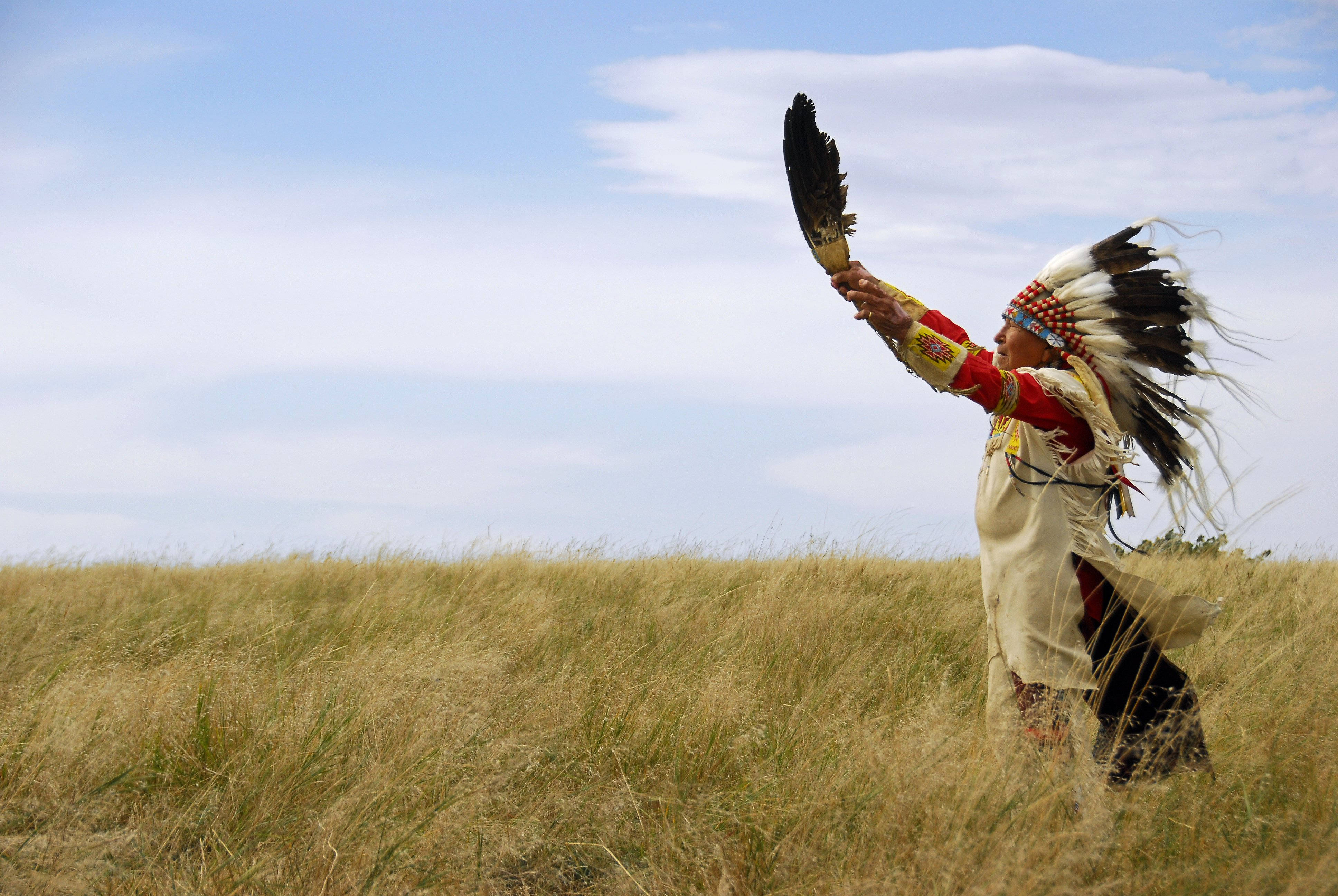 Native American 桌布 壁紙 — 一位身穿傳統服飾的美洲原住民站在高高的金色草地上，在明亮的藍天下，以儀式性的姿態舉起一根飾有羽毛的杖。