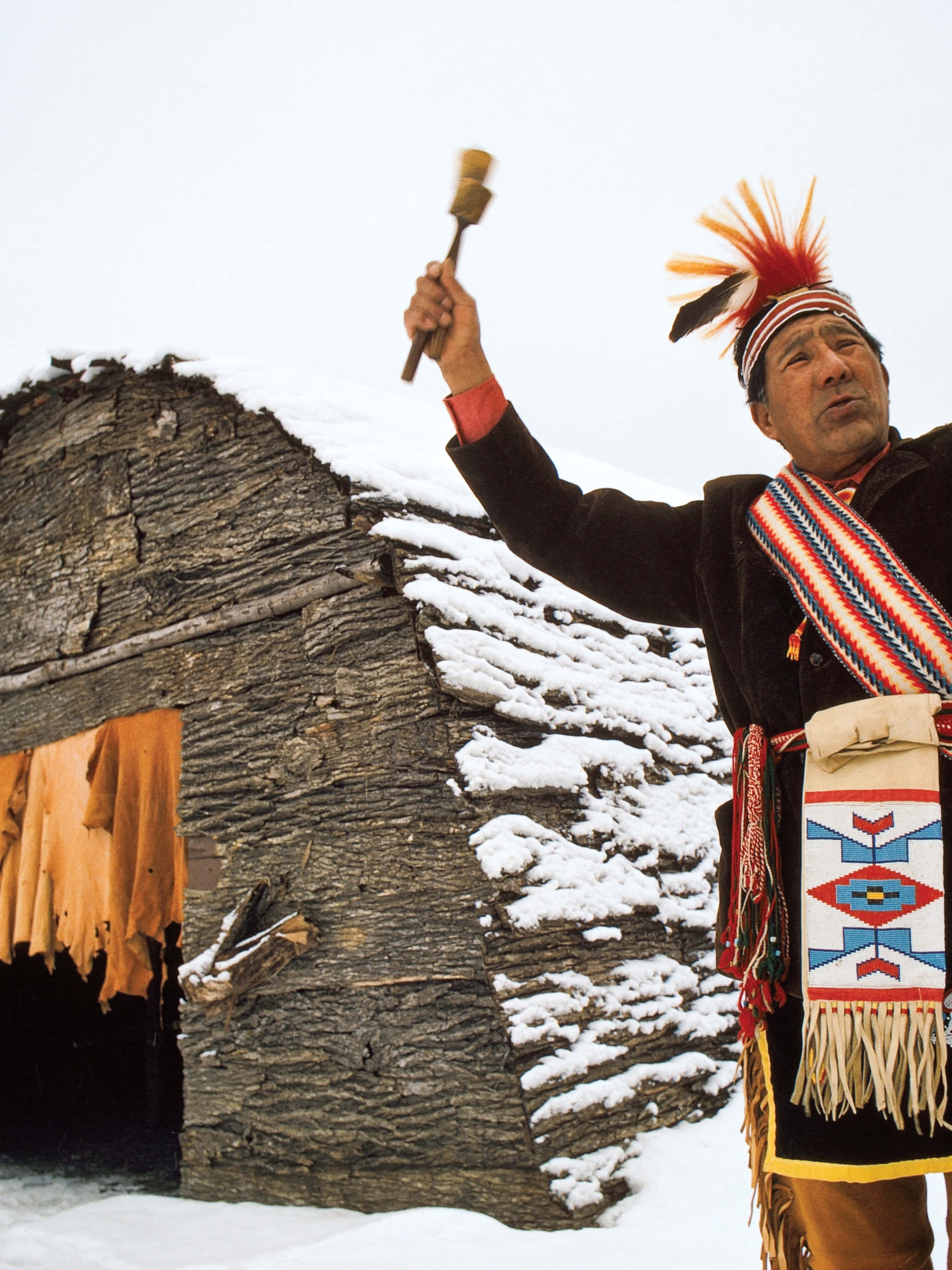 Native American 桌布 壁紙 — 一位身穿傳統服飾的長者在嚴冬的景色中，在灑滿白雪的石屋前舉起儀式用的搖鈴。