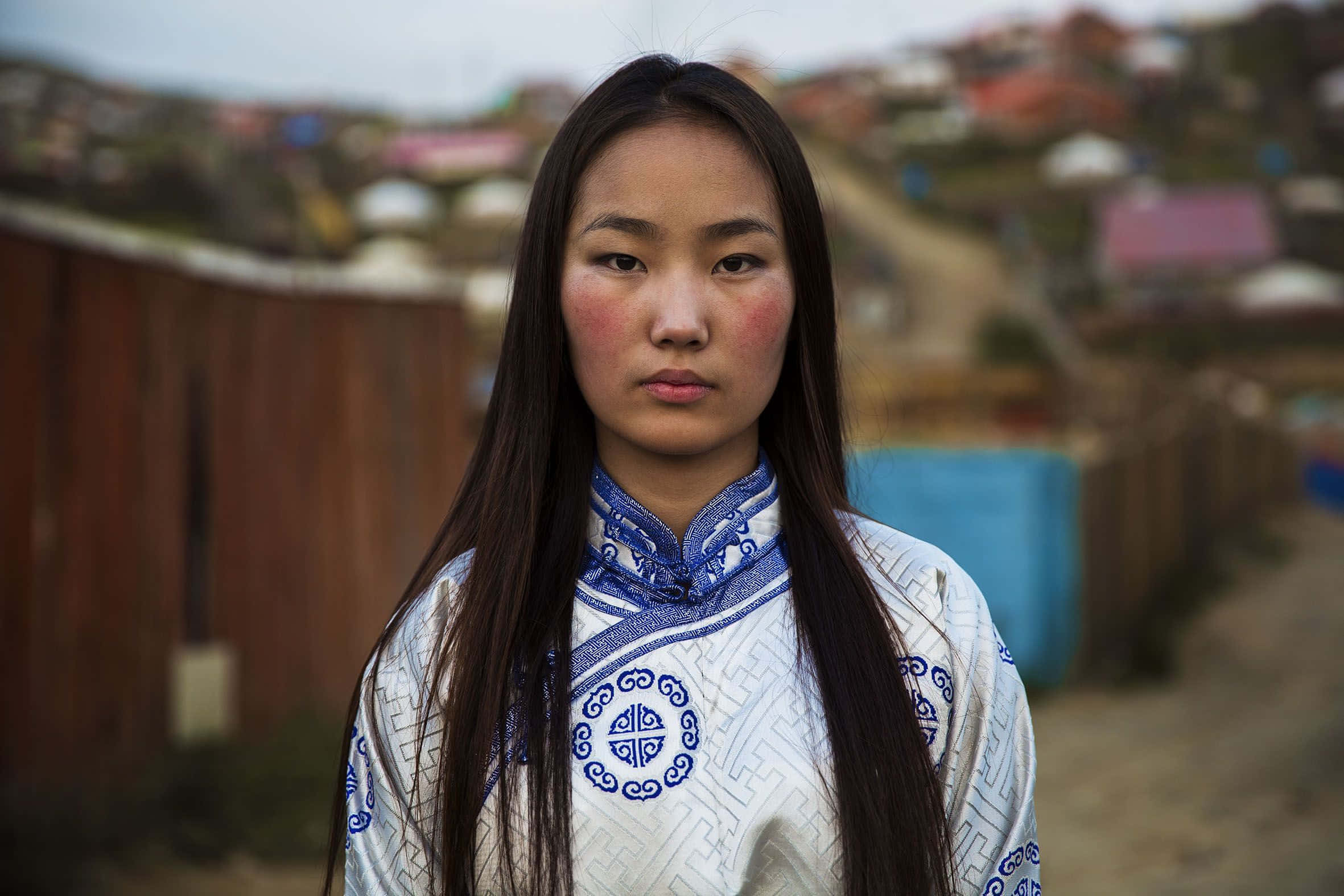 Woman 桌布 壁紙 — 一位身穿傳統藍白刺繡服飾的年輕女子，在模糊的山坡村莊背景下莊嚴地站立。