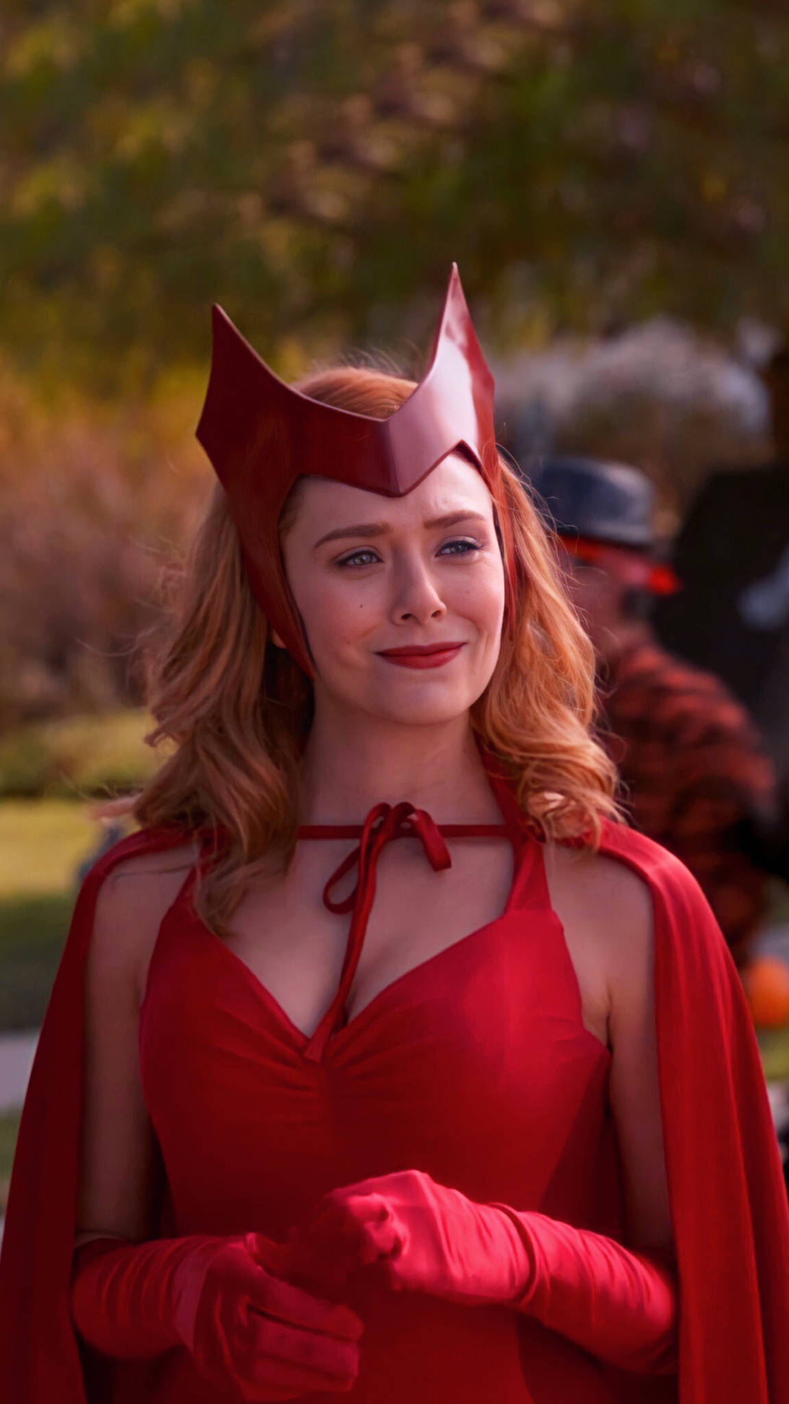 Wanda 桌布 壁紙 — 一位身穿全套紅色女巫服裝、戴著尖帽和披風的女性在戶外溫柔微笑。