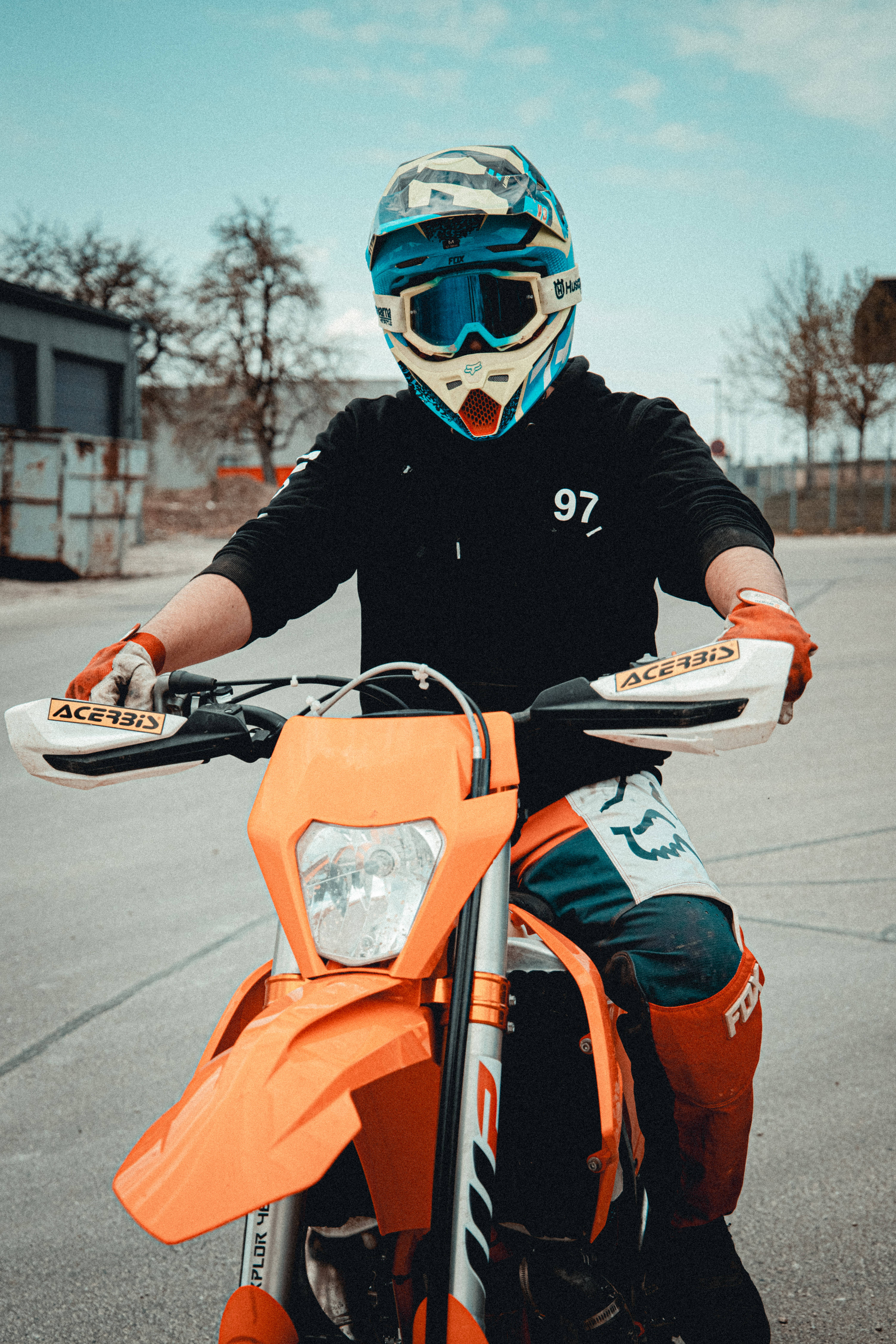 Ktm Bike 桌布 壁紙 — 一位身穿全套裝備的越野摩托車手，在晴朗天空下的柏油地上，握著一輛亮橙色越野車的把手。
