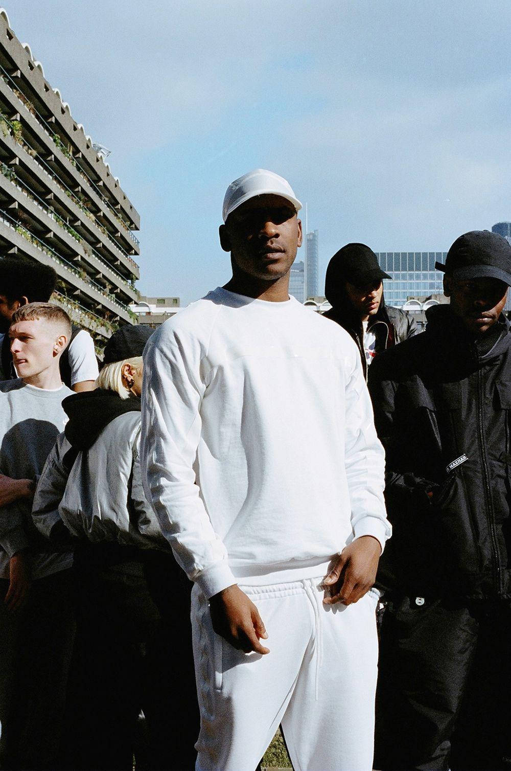 Skepta 桌布 壁紙 — 一位身穿全白衣服的年輕男子自信地站在戶外，身後是城市建築，其他人聚集在周圍。