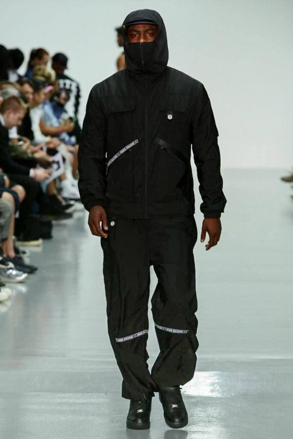 Skepta 桌布 壁紙 — 一位身穿全黑連帽服裝的模特兒自信地走在極簡主義的伸展台上，觀眾坐在兩旁。
