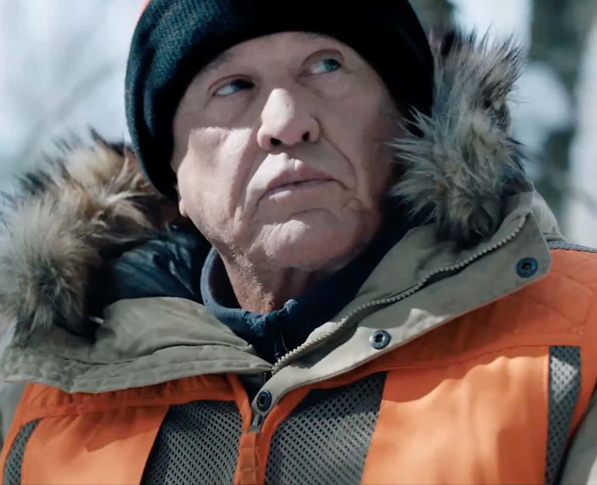 Tom Berenger 桌布 壁紙 — 一位身穿冬季外套和毛帽的長者，在雪景背景下帶著嚴肅的表情向上凝視。