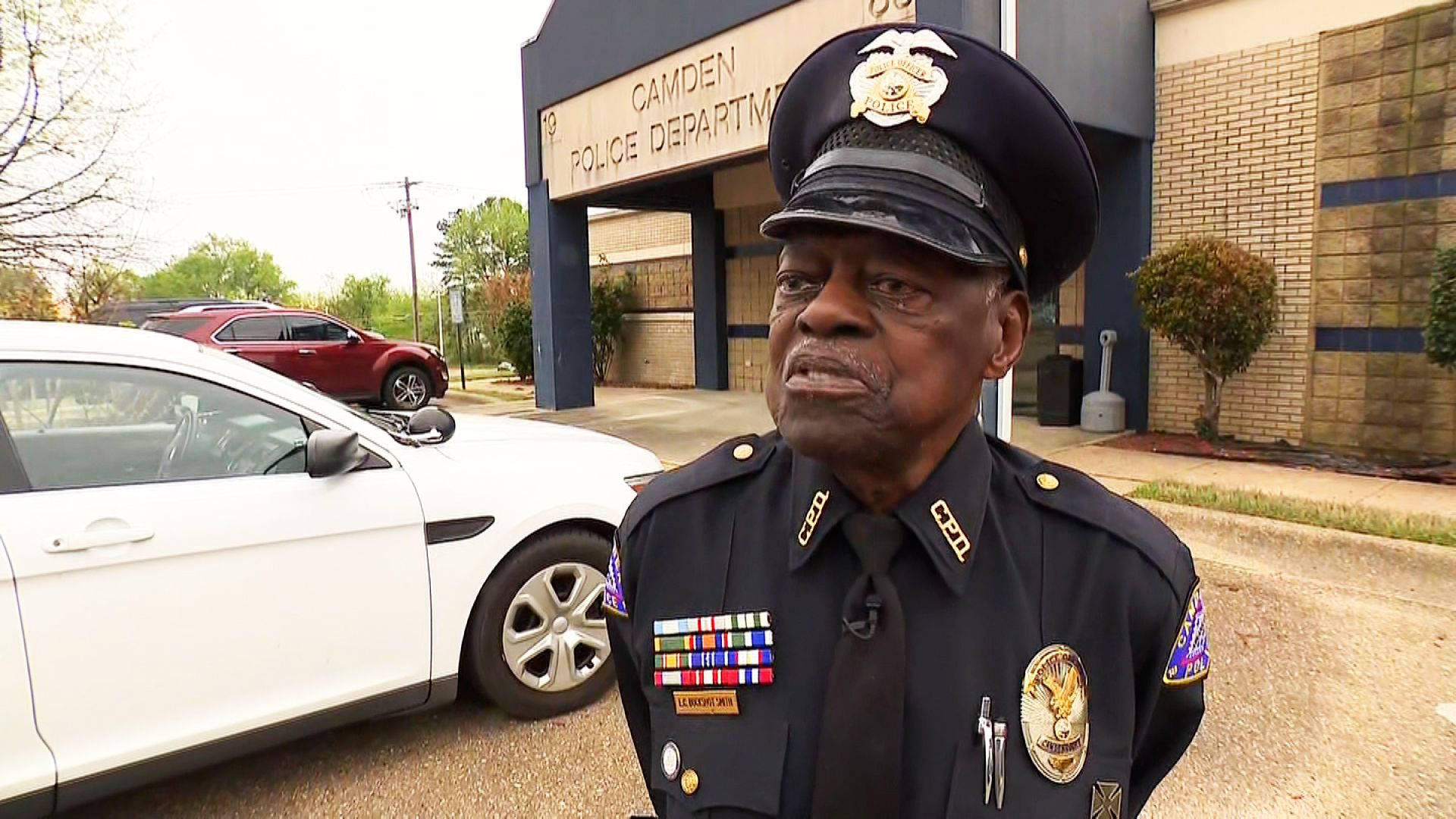 job 桌布 壁紙 — 一位身穿制服的高級警察局長站在卡姆登警察局外，附近停著一輛白色汽車。