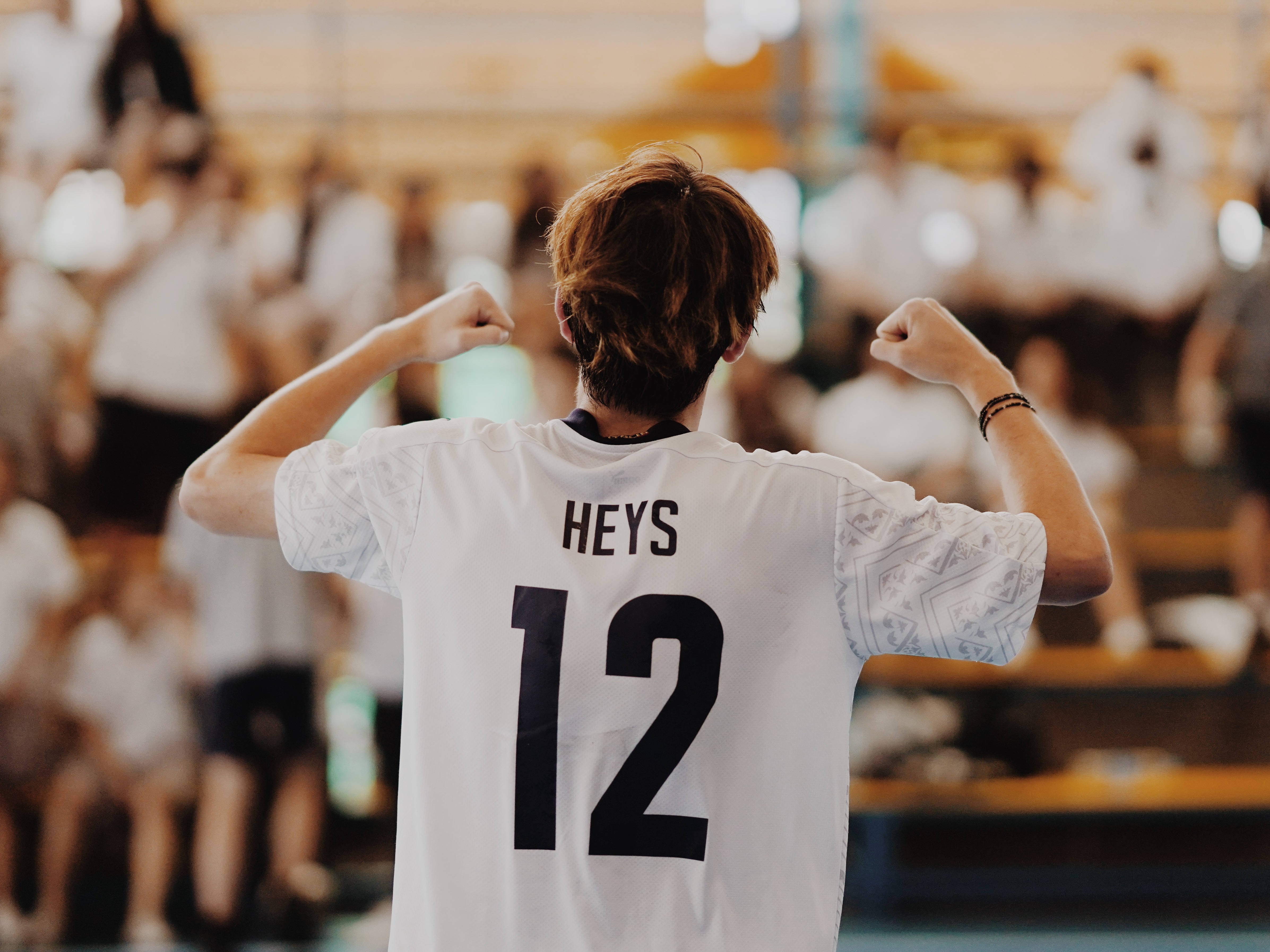 排球 桌布 壁紙 — 一位身穿印有HEYS 12白色球衣的年輕運動員，在體育館內面對模糊的人群，勝利地彎曲雙臂。
