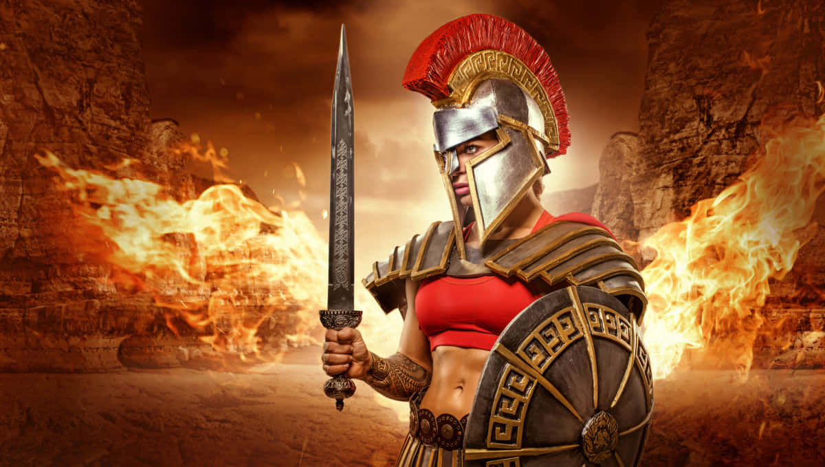 Gladiator 桌布 壁紙 — 一位身穿古希臘盔甲的兇猛女戰士，在岩石峽谷中手持劍和盾牌，身後火焰噴發，她挑釁地站立著。