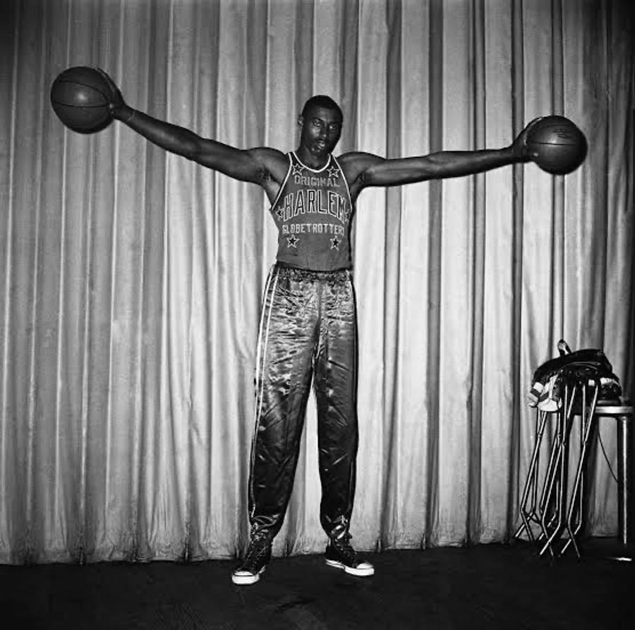 籃球 桌布 壁紙 — 一位身穿哈林環球旅行者球衣的高大籃球運動員，在垂簾背景下雙臂張開，每隻手各持一個球。