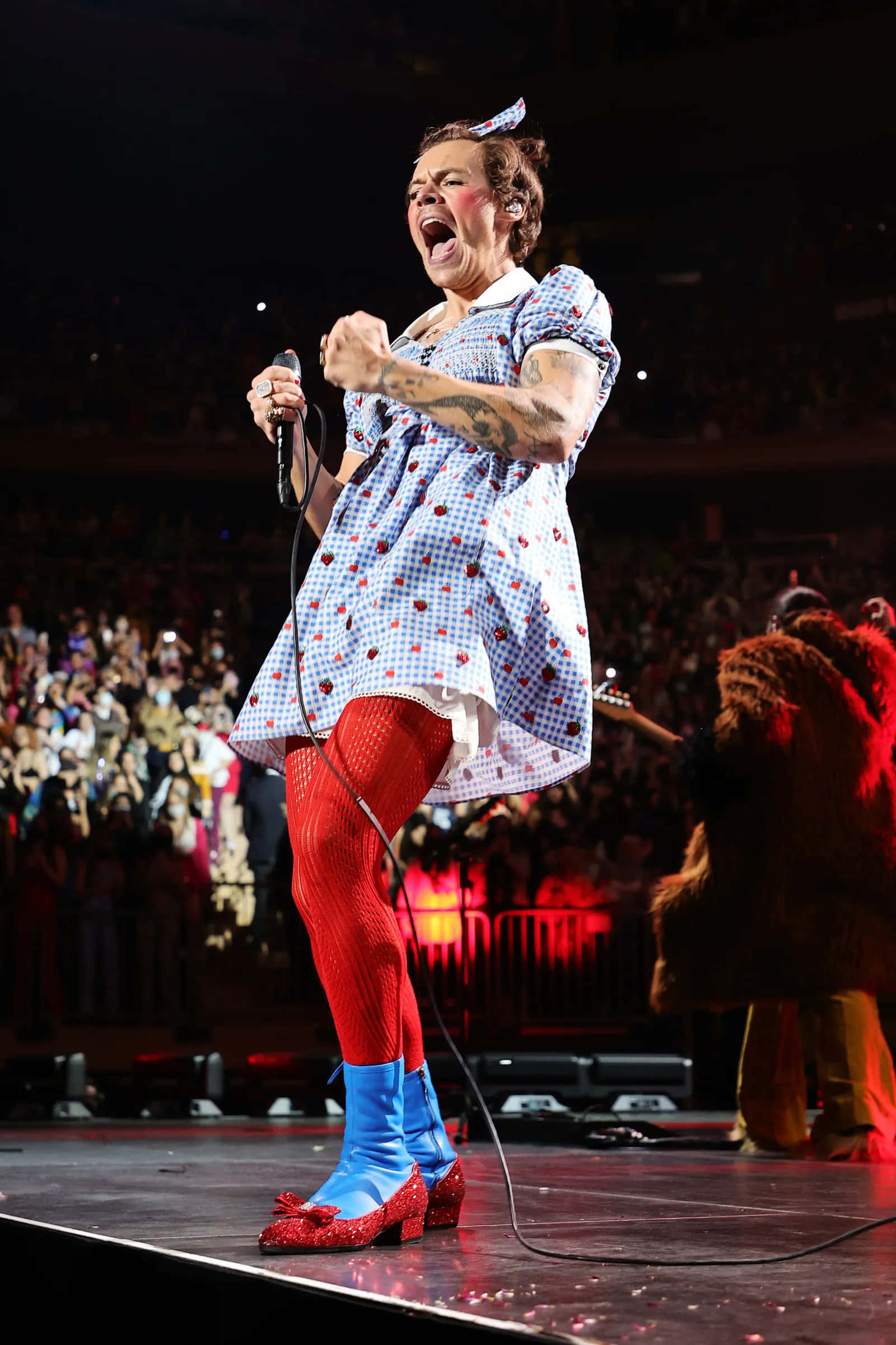 Harry Styles 2021 桌布 壁紙 — 一位身穿圓點裙和紅色緊身褲的歌手在燈光璀璨的舞台上對著麥克風高歌，台下觀眾歡呼雷動。