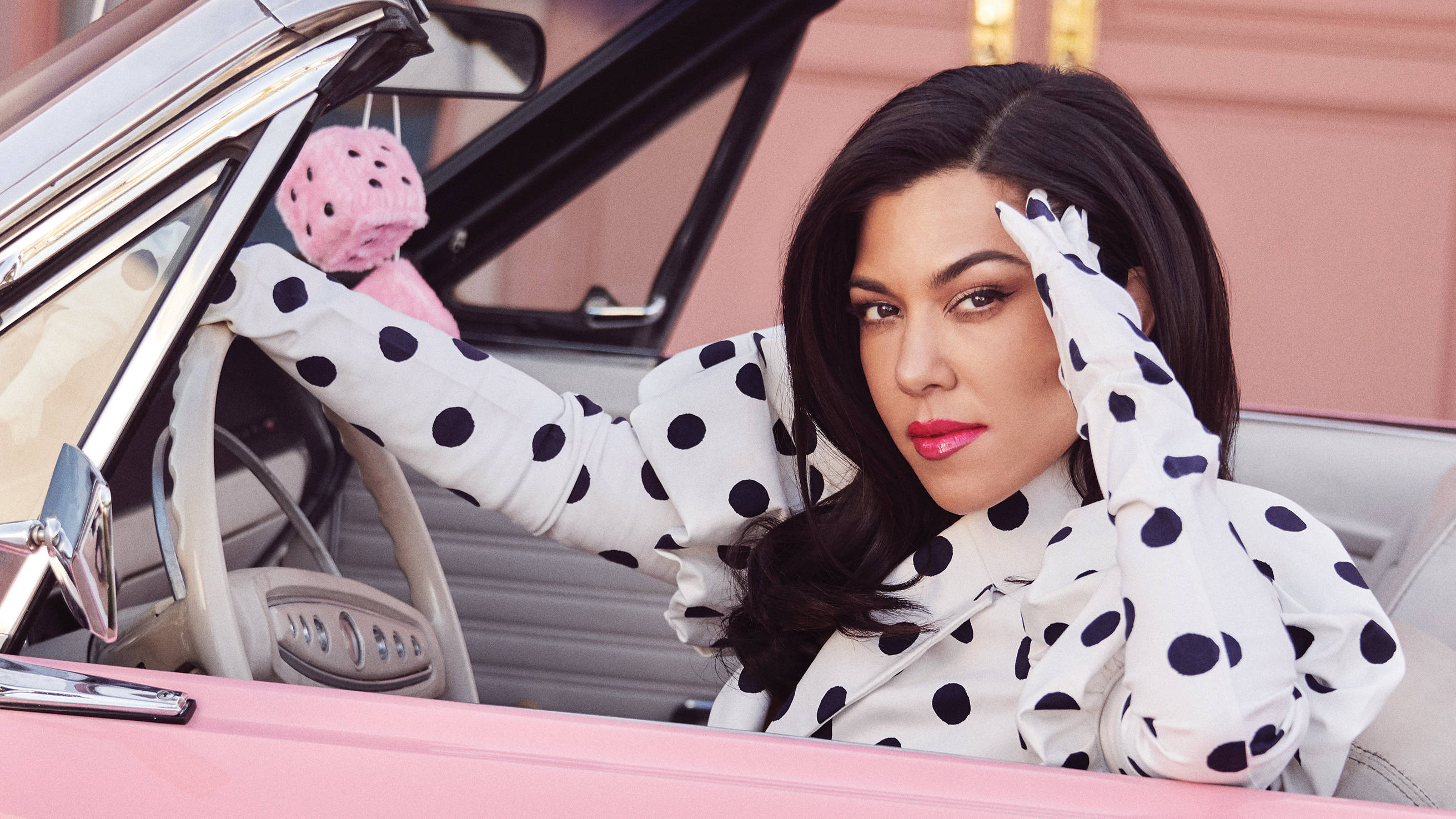 Kourtney 桌布 壁紙 — 一位身穿圓點襯衫的女性從一輛粉紅色敞篷車中探出身子，後視鏡上掛著一個毛絨草莓。