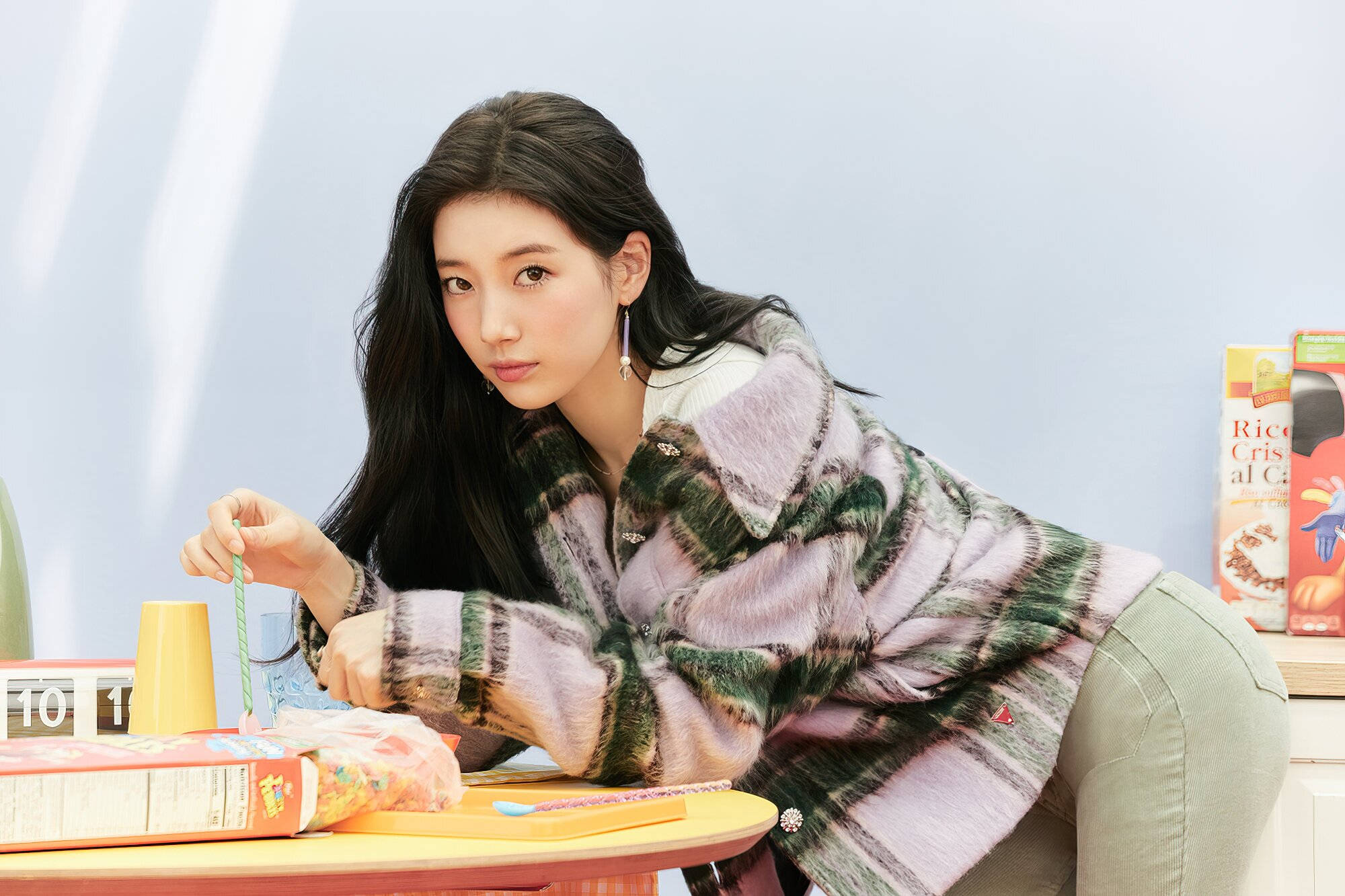 bae 桌布 壁紙 — 一位身穿圖案外套的年輕女子俯身靠近放有穀物盒和吸管的桌子，以柔和的表情凝視鏡頭。
