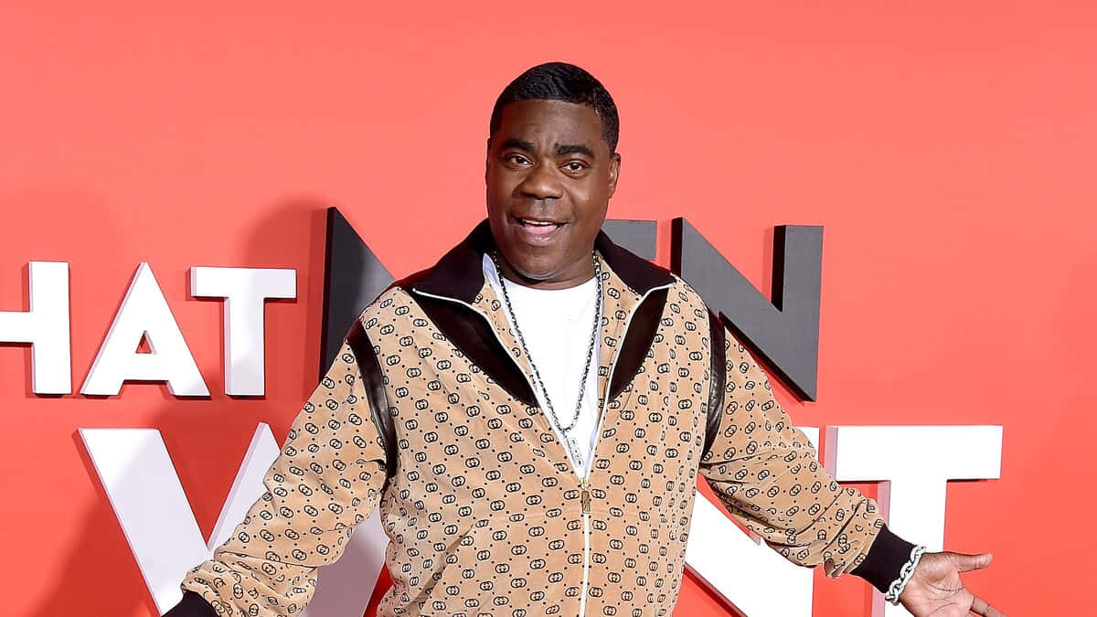Tracy Morgan 桌布 壁紙 — 一位身穿圖案米色外套的男子，在帶有白色字母的鮮豔橙色背景前自信地微笑。