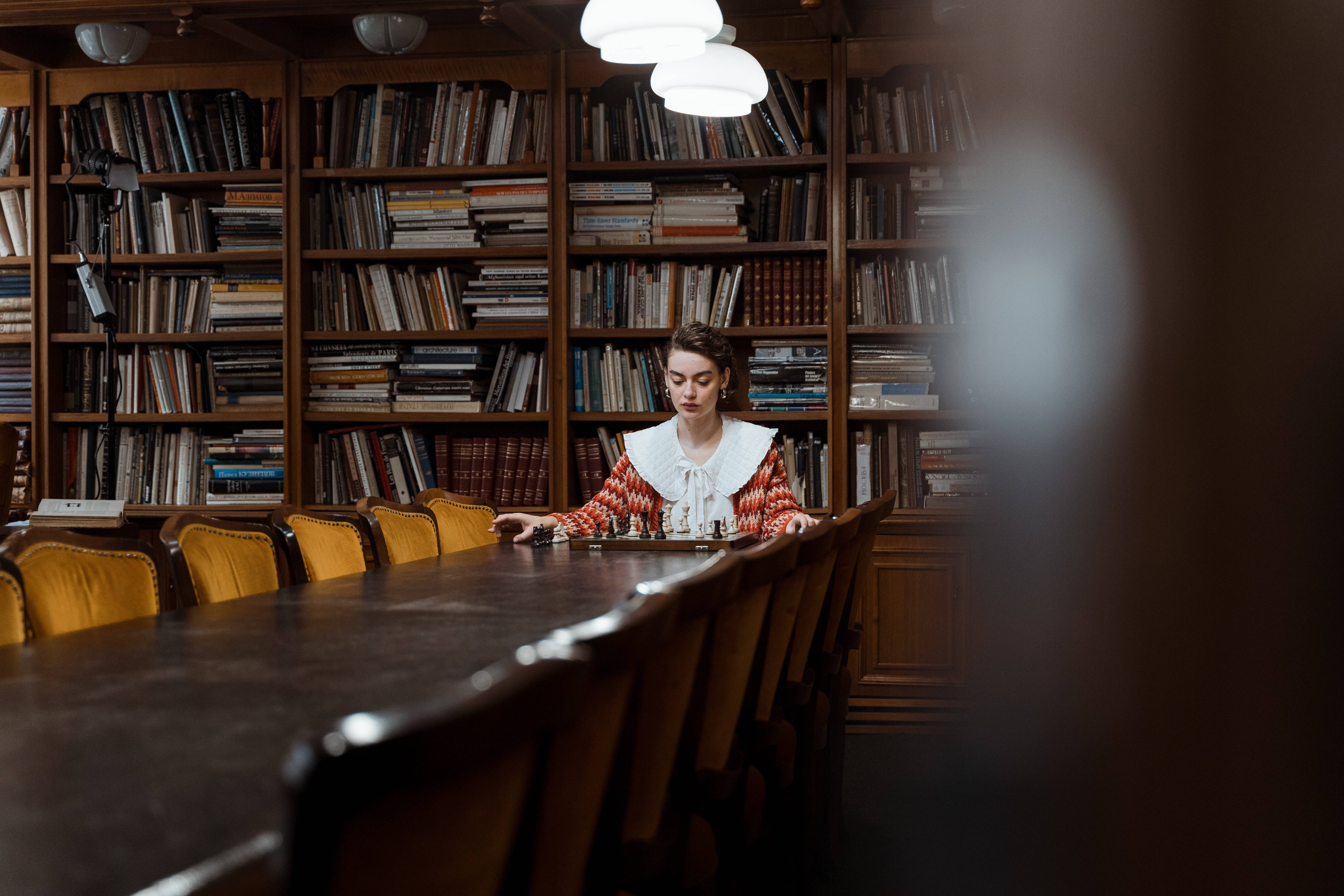 暗黑學院風 桌布 壁紙 — 一位身穿圖案襯衫的女性在燈光昏暗的圖書館中，獨自在一張長木桌上玩西洋棋，周圍是高聳的書架。