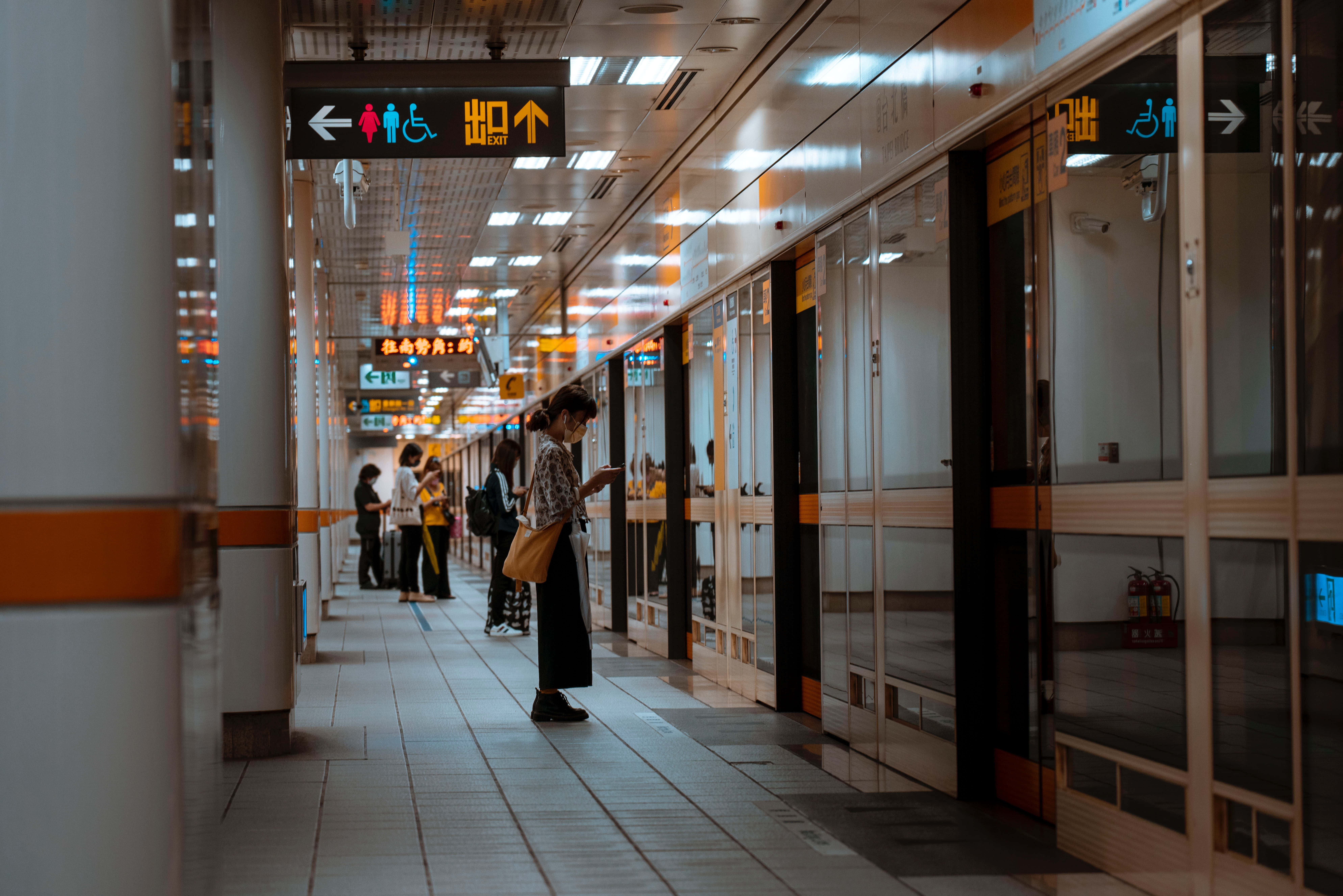 Waiting 桌布 壁紙 — 一位身穿圖案襯衫的女性獨自站在現代化的地鐵月台上，頭頂明亮的燈光，遠處有其他通勤者。