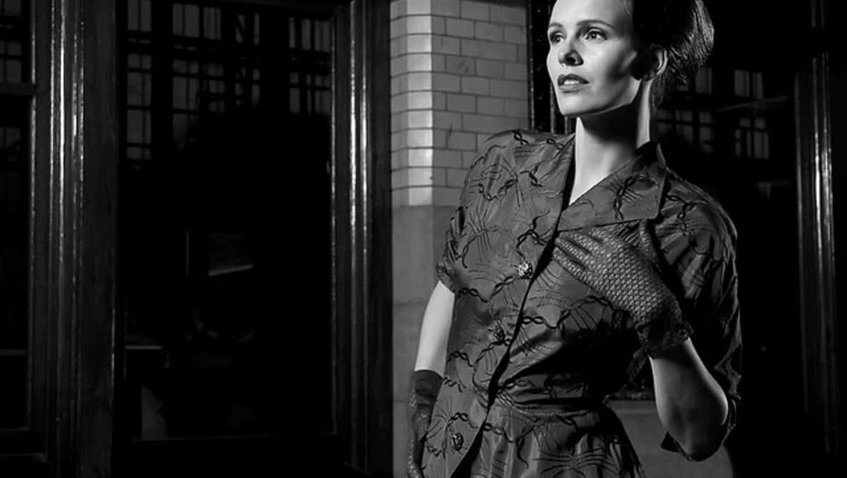 Film Noir 桌布 壁紙 — 一位身穿圖案連衣裙的女性站在光線昏暗的門口，她的臉被戲劇性的明暗對比光照亮。
