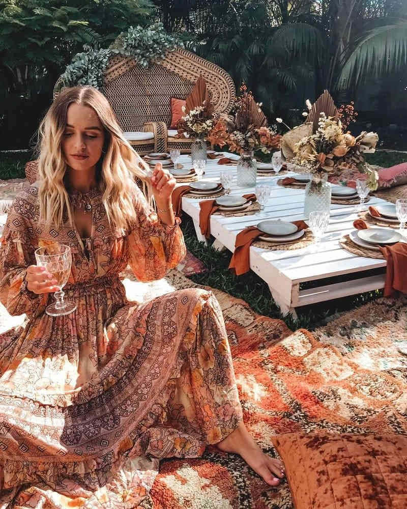 美學波希米亞 桌布 壁紙 — 一位身穿圖案連衣裙的女性赤腳坐在色彩繽紛的地毯上，旁邊是一張白色木桌，擺設成波希米亞風格的戶外聚會，上方是茂密的綠植。