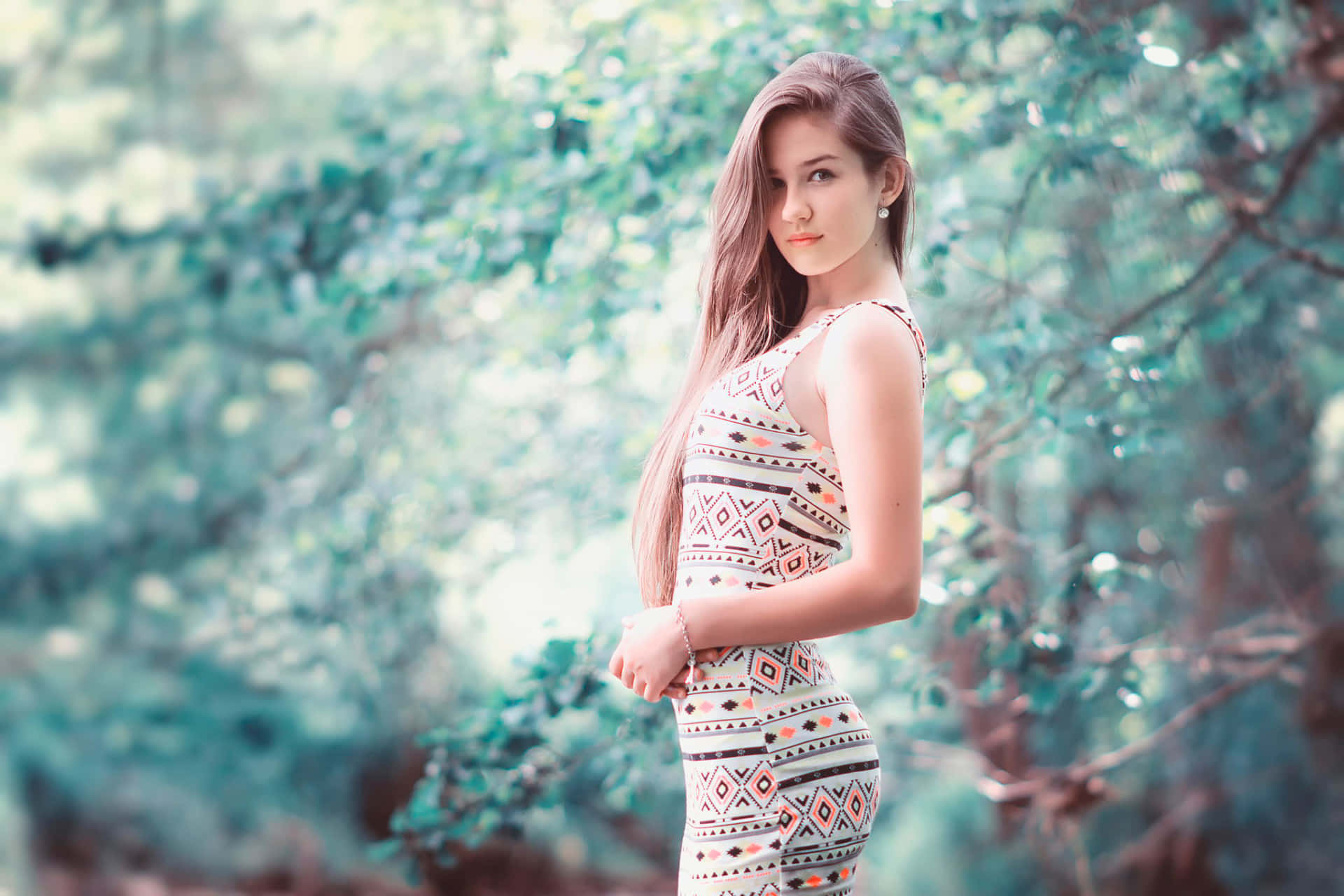 美女 桌布 壁紙 — 一位身穿圖案連衣裙的年輕女子優雅地站在陽光明媚的森林中，身後是柔和的散景樹葉。