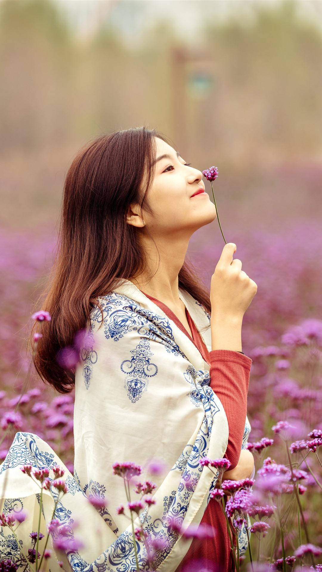 美女 桌布 壁紙 — 一位身穿圖案長袍的年輕女性，在一片盛開的紫色花朵田野中，輕輕聞著一朵紫色的花。