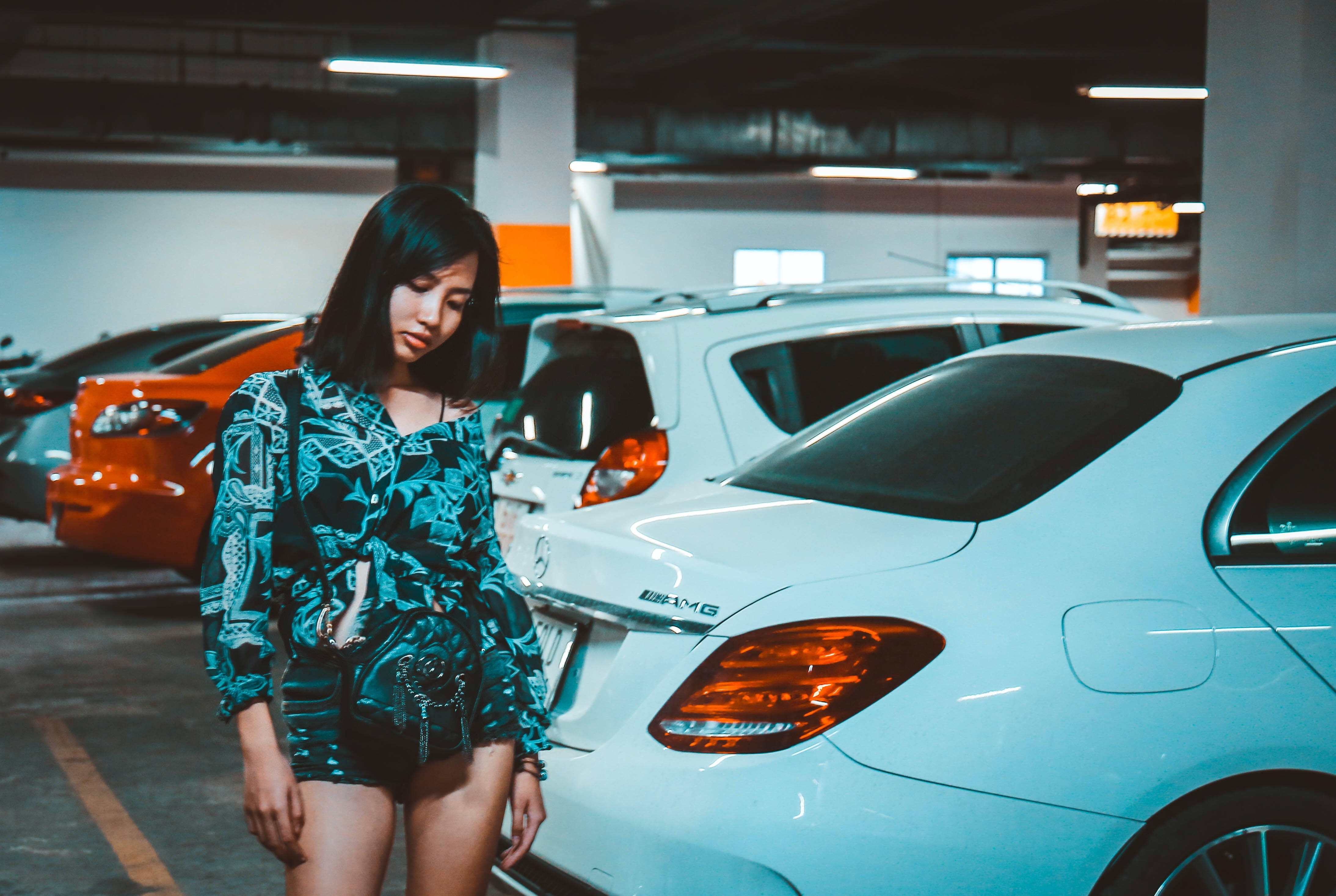 Woman 桌布 壁紙 — 一位身穿圖案青綠色套裝的年輕女性，站在昏暗的地下停車場中一輛白色Mercedes AMG旁。