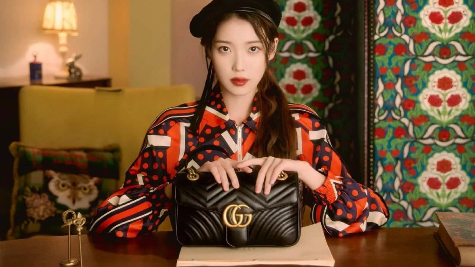 Gucci 桌布 壁紙 — 一位身穿大膽圖案襯衫和貝雷帽的女性，在木桌上拿著一個黑色絎縫Gucci手袋，背景是鮮豔的花卉壁紙。
