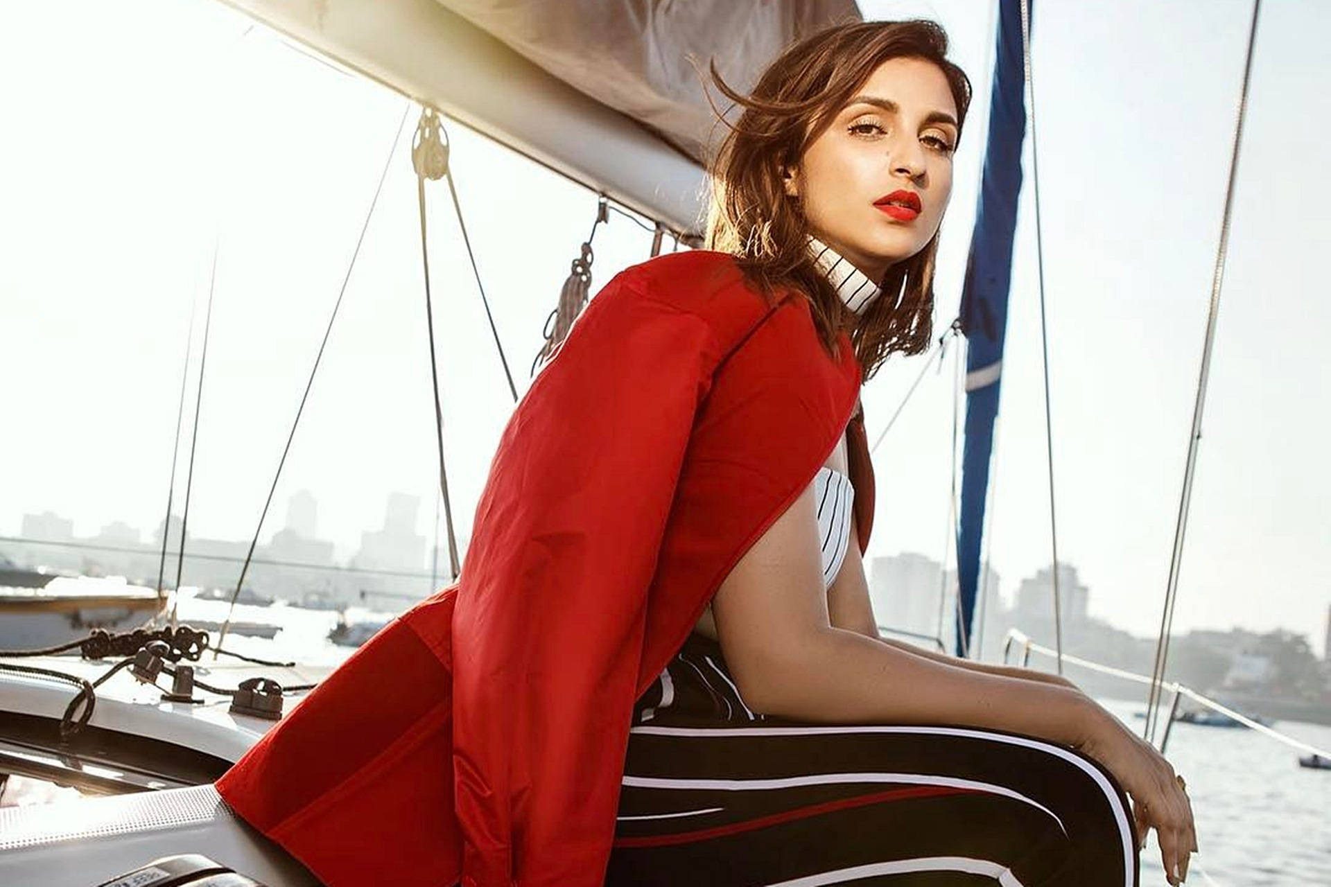 Parineeti 桌布 壁紙 — 一位身穿大膽紅色外套的女性自信地坐在帆船上，背景是朦朧的城市天際線。
