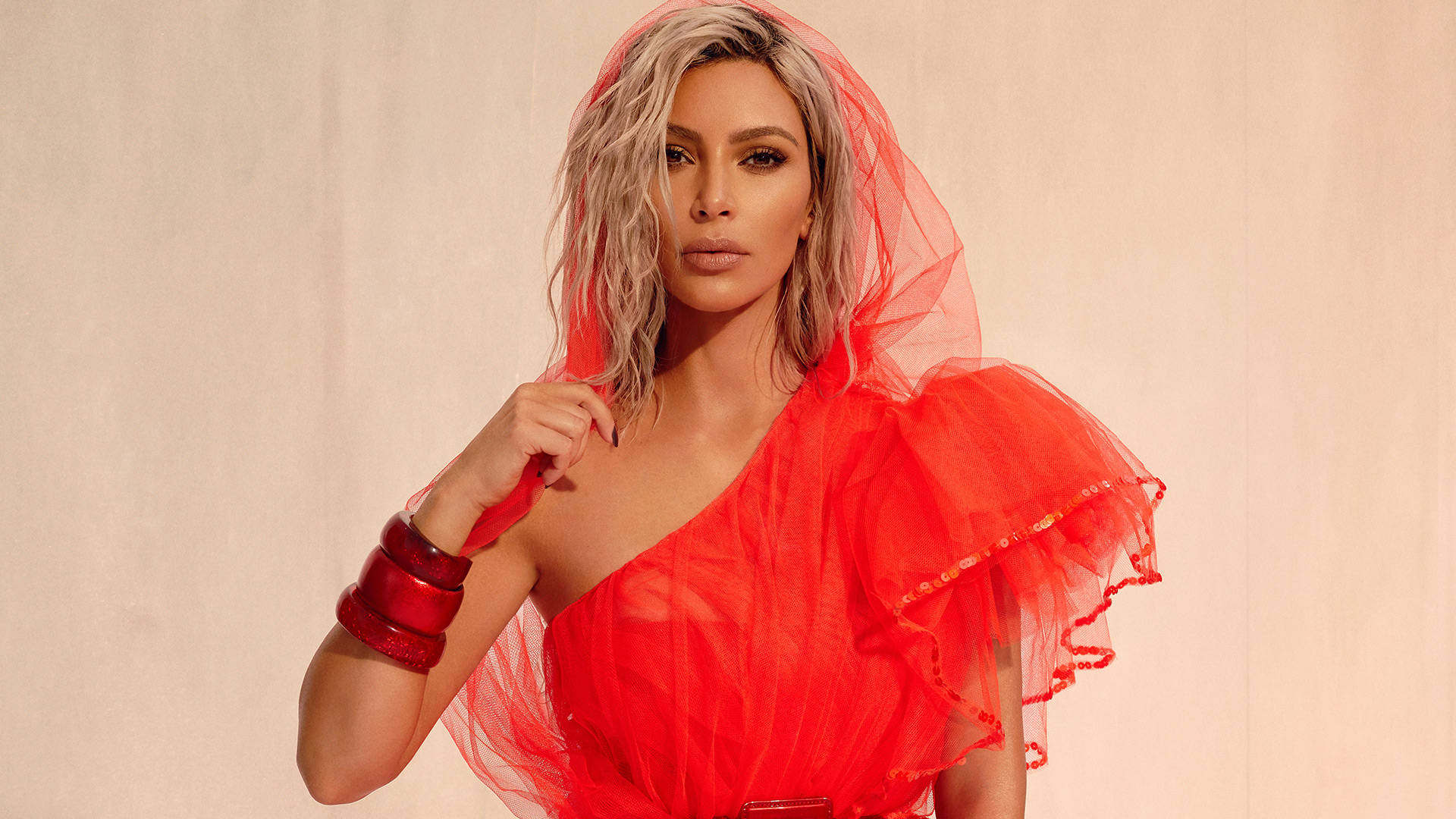 Kim Kardashian 桌布 壁紙 — 一位身穿大膽紅色薄紗長裙和相配頭飾的女性，在柔和的米色背景前自信地擺姿勢。