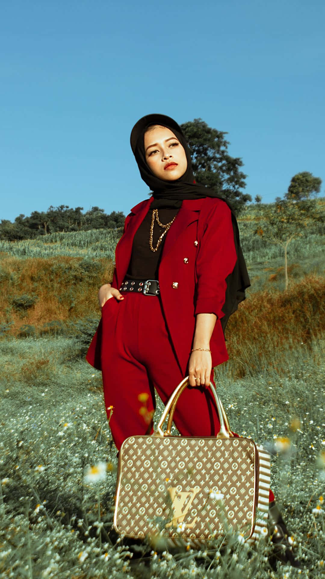 louis vuitton phone 桌布 壁紙 — 一位身穿大膽紅色西裝和黑色希賈布的女性，自信地站在陽光明媚的野花田中，手持一款圖案設計師手袋。