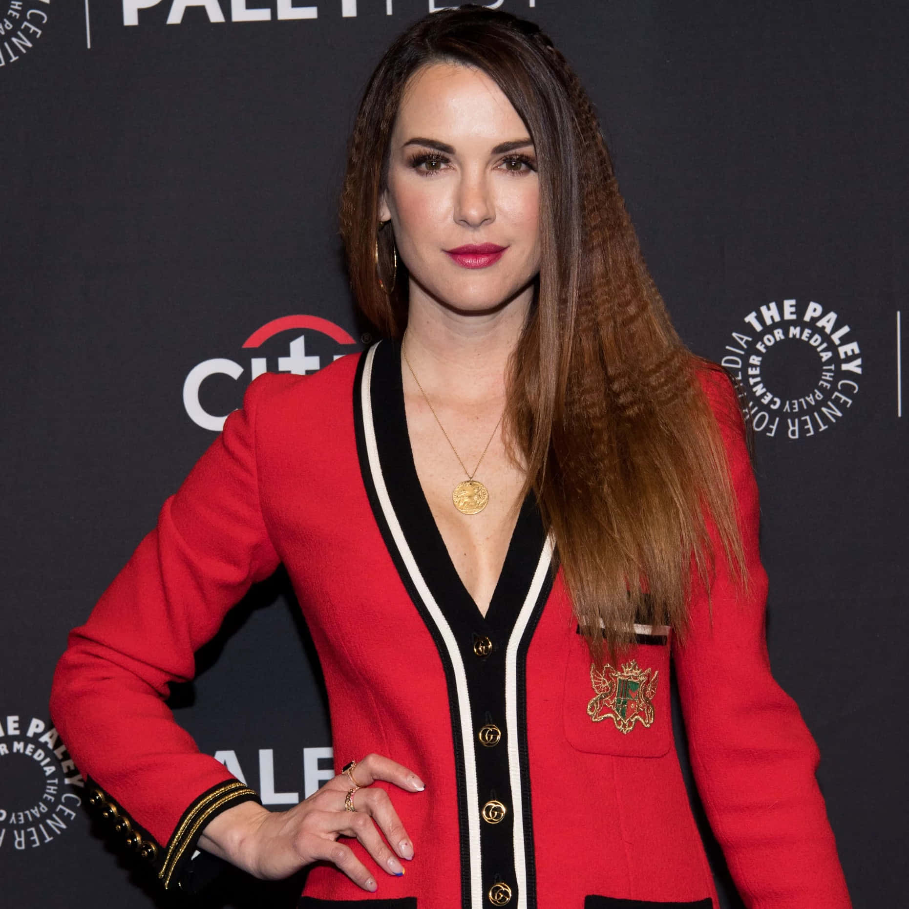 Danneel Ackles 桌布 壁紙 — 一位身穿大膽紅色西裝外套、帶有黑白飾邊的女性，在品牌背景的媒體活動中自信地擺姿勢。