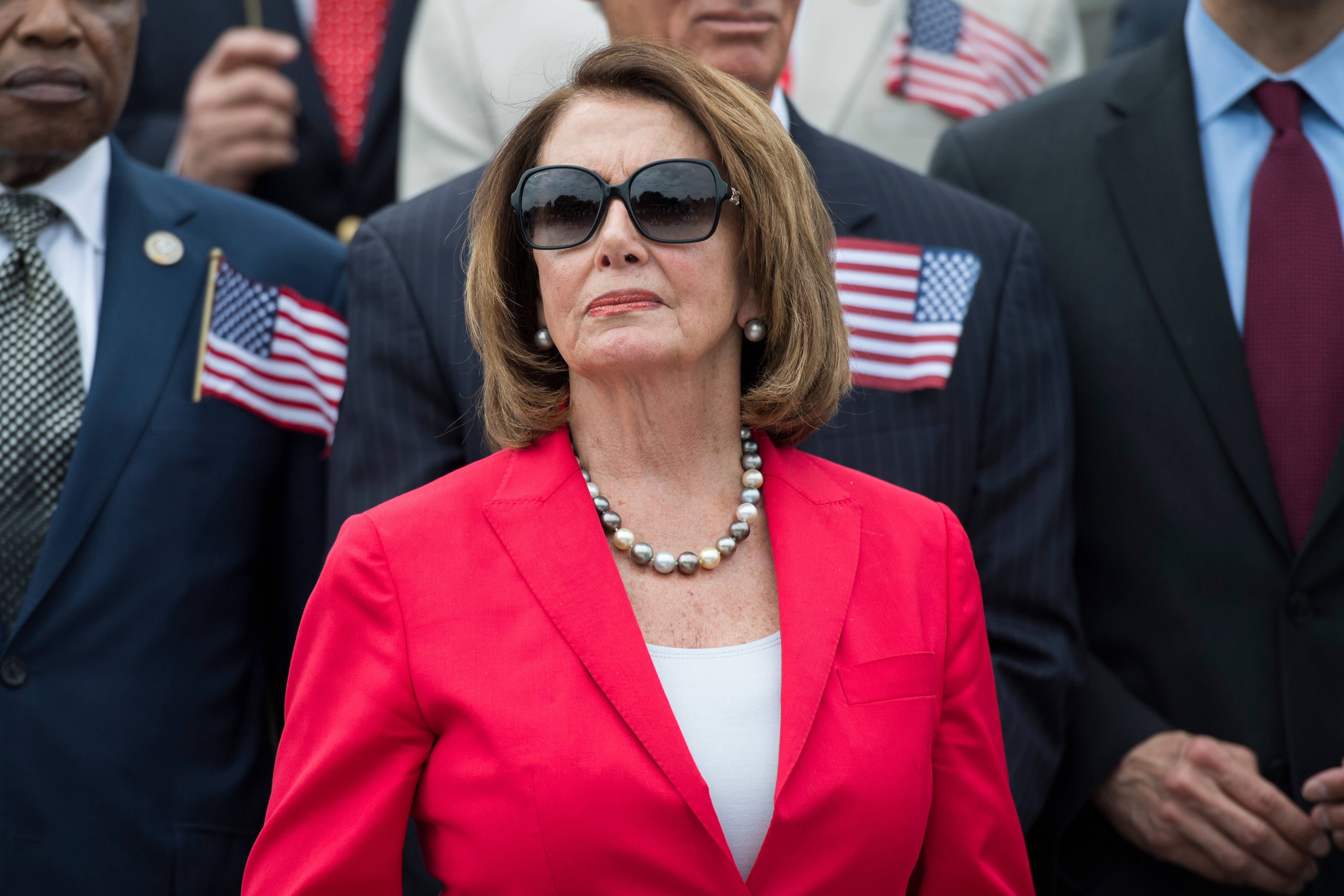 Nancy Pelosi 桌布 壁紙 — 一位身穿大膽紅色西裝外套和太陽眼鏡的女性，莊嚴地站在一群穿著西裝、翻領上別著美國國旗的男士中間。