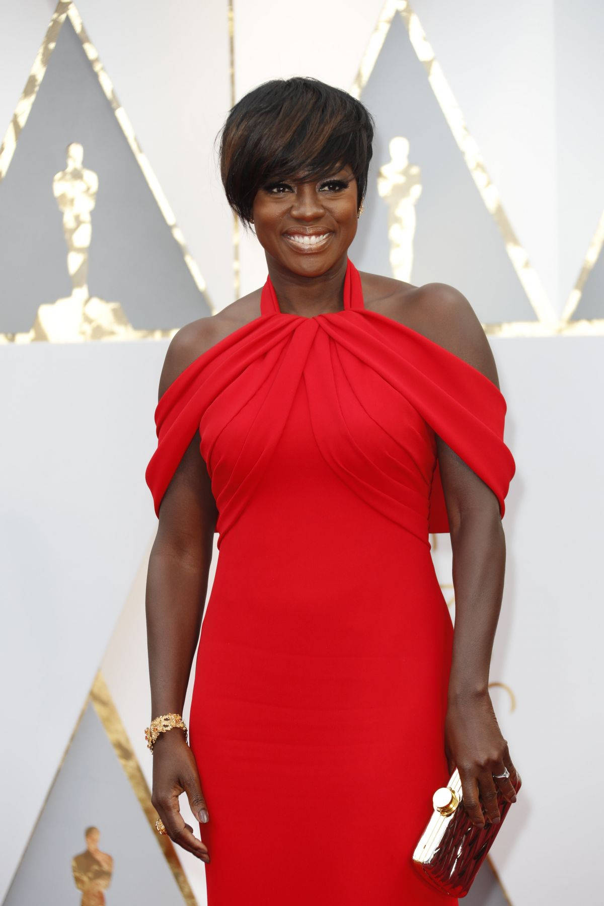 oscars 桌布 壁紙 — 一位身穿大膽紅色露肩禮服的耀眼女性，在華麗的紅地毯上燦爛微笑，背景是金色的奧斯卡小金人。