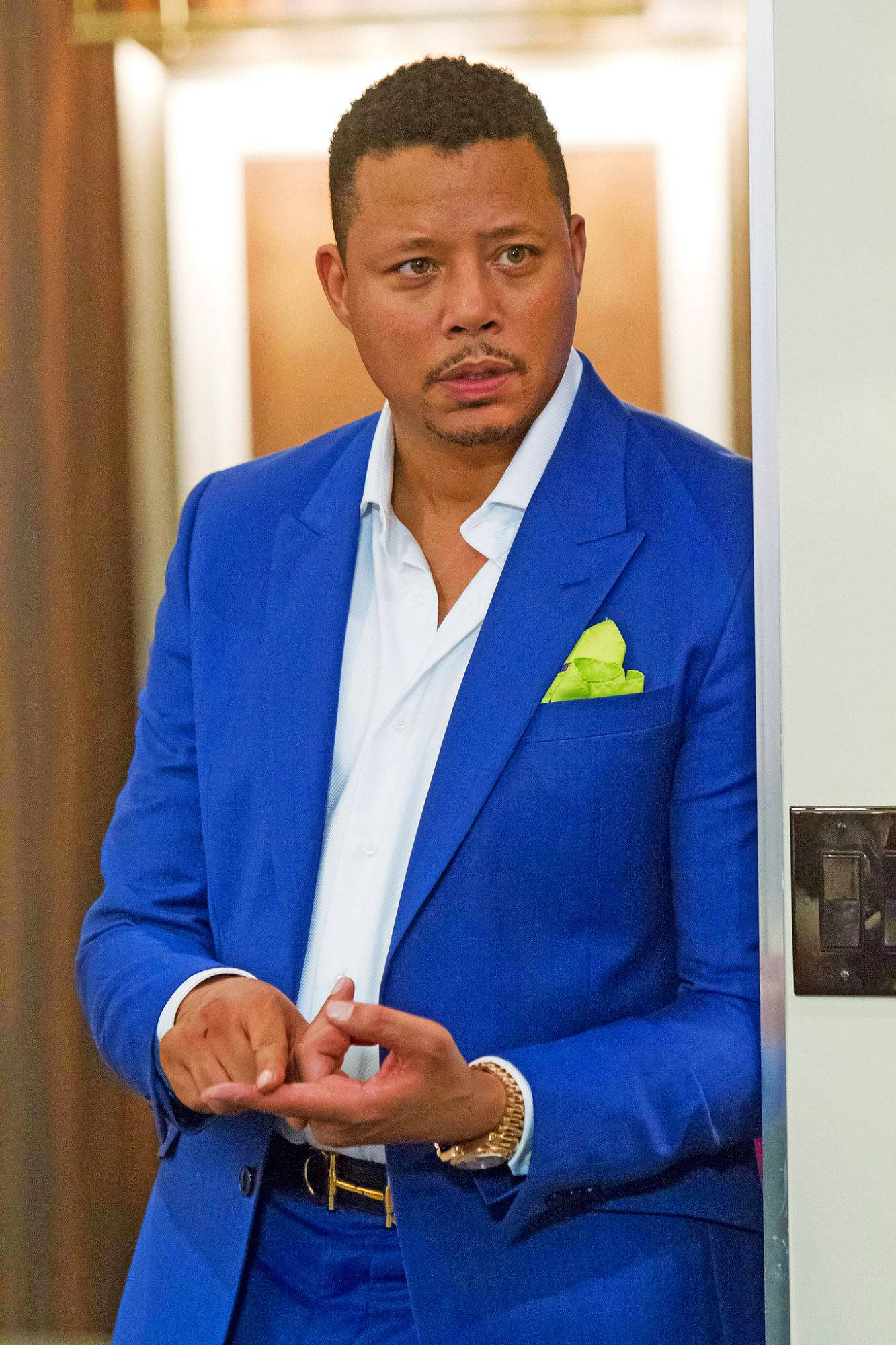 Entertainment 桌布 壁紙 — 一位身穿大膽藍色西裝、搭配萊姆綠色口袋方巾的男子站在門口，雙手緊握，專注地向前凝視。