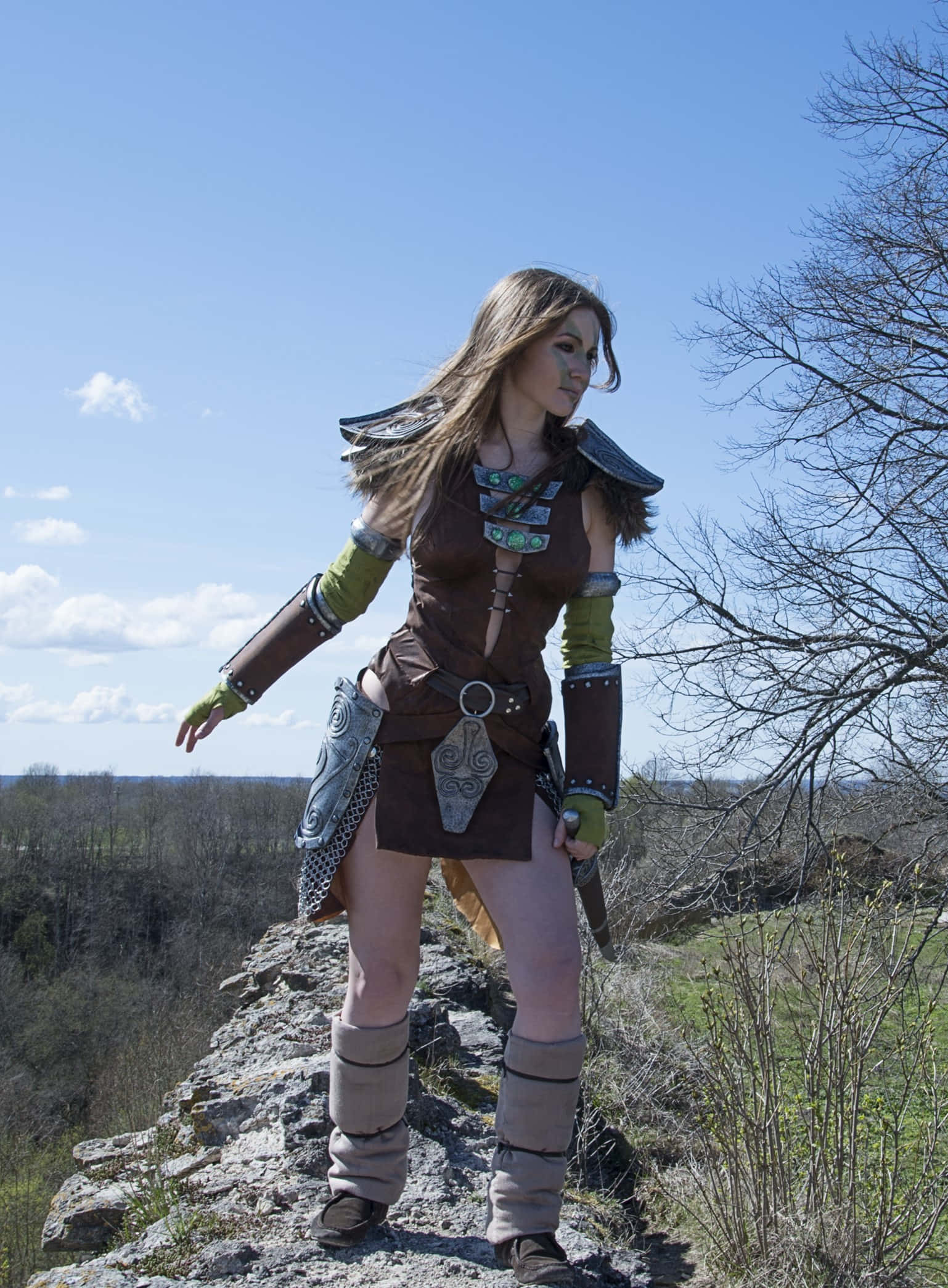 Aela The Huntress 桌布 壁紙 — 一位身穿奇幻盔甲的女性自信地站在岩石懸崖邊緣，頭頂是明亮藍天，點綴著稀疏雲朵。