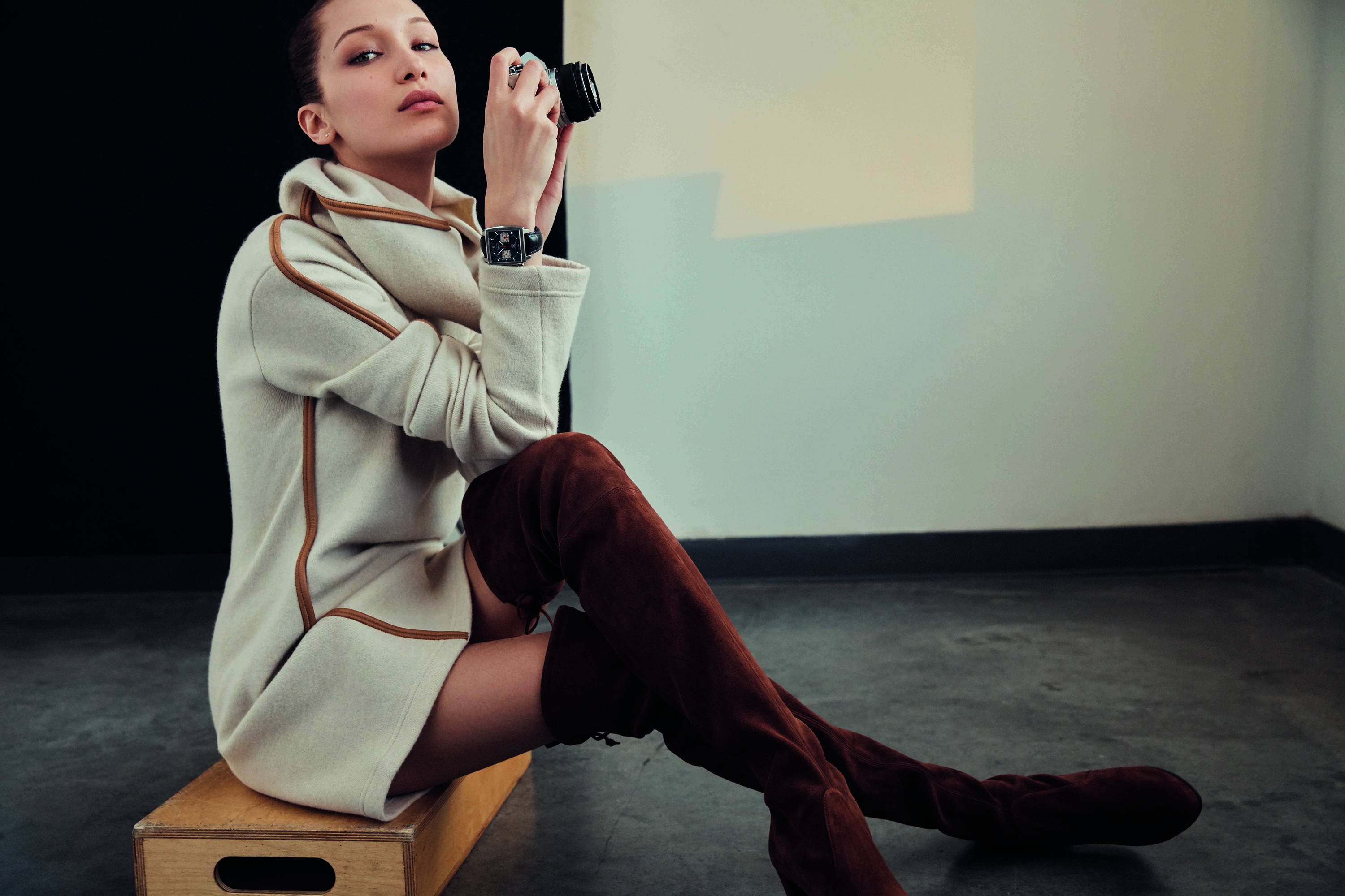 Bella Hadid 桌布 壁紙 — 一位身穿奶油色外套和酒紅色靴子的女性坐在木箱上，在簡約的工作室中手持相機，光線柔和。