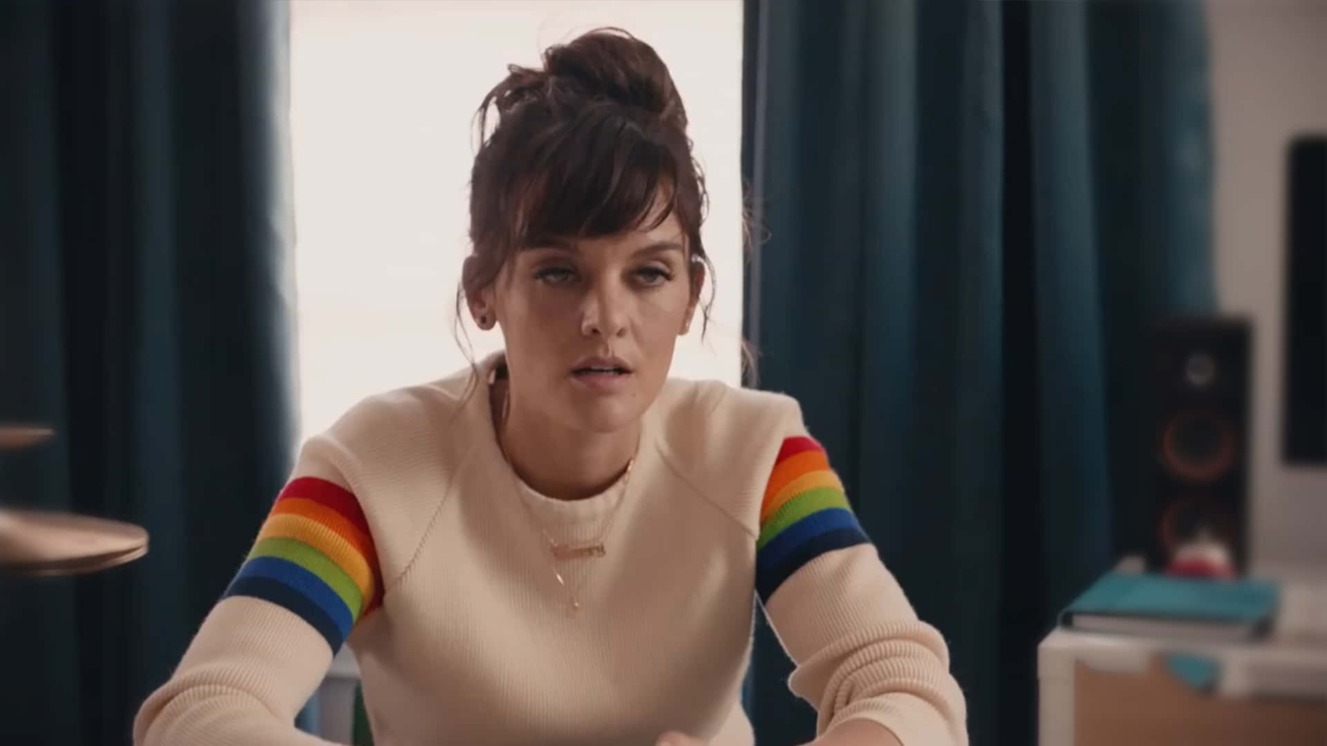 Frankie Shaw 桌布 壁紙 — 一位身穿奶油色毛衣、袖子上有彩虹條紋的女性坐在室內，身後有深色窗簾，她專注地向前看。