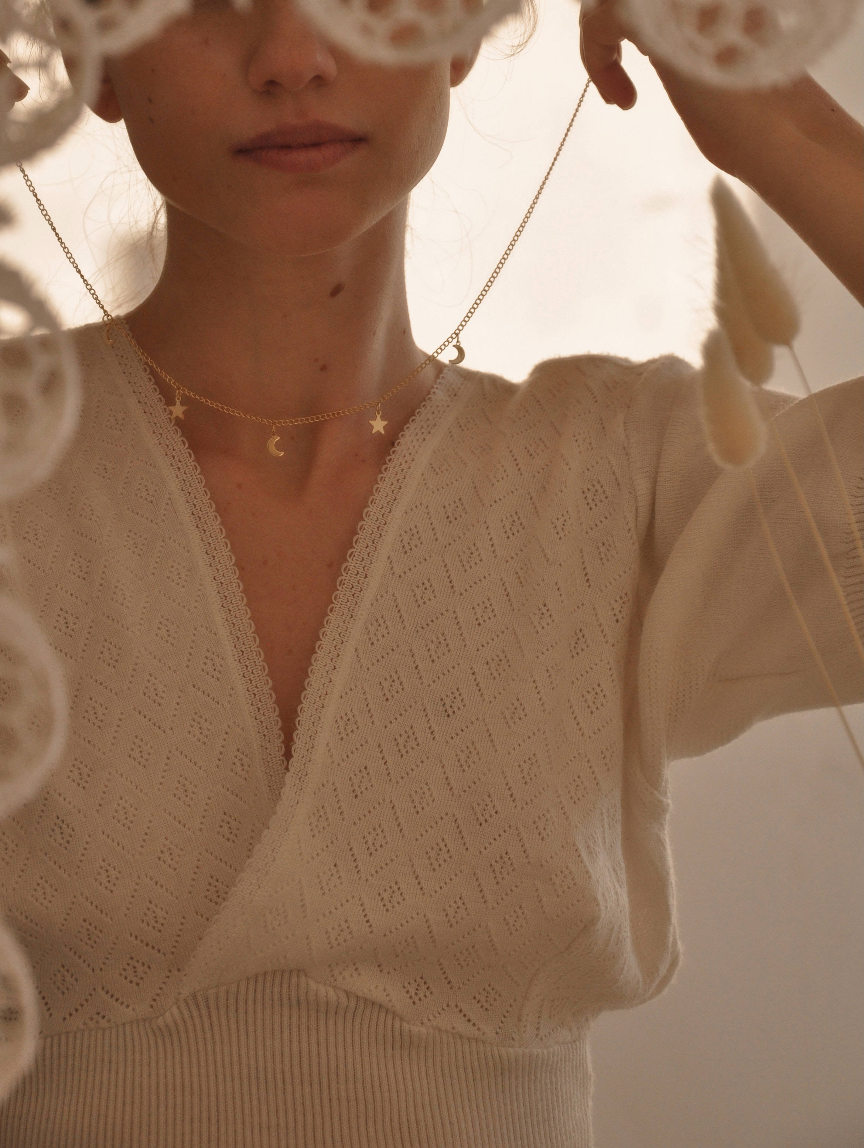Gold Jewellery 桌布 壁紙 — 一位身穿奶油色針織上衣的女性，在柔和的金色光線中手持精緻的白色蕾絲，佩戴著星月項鍊。