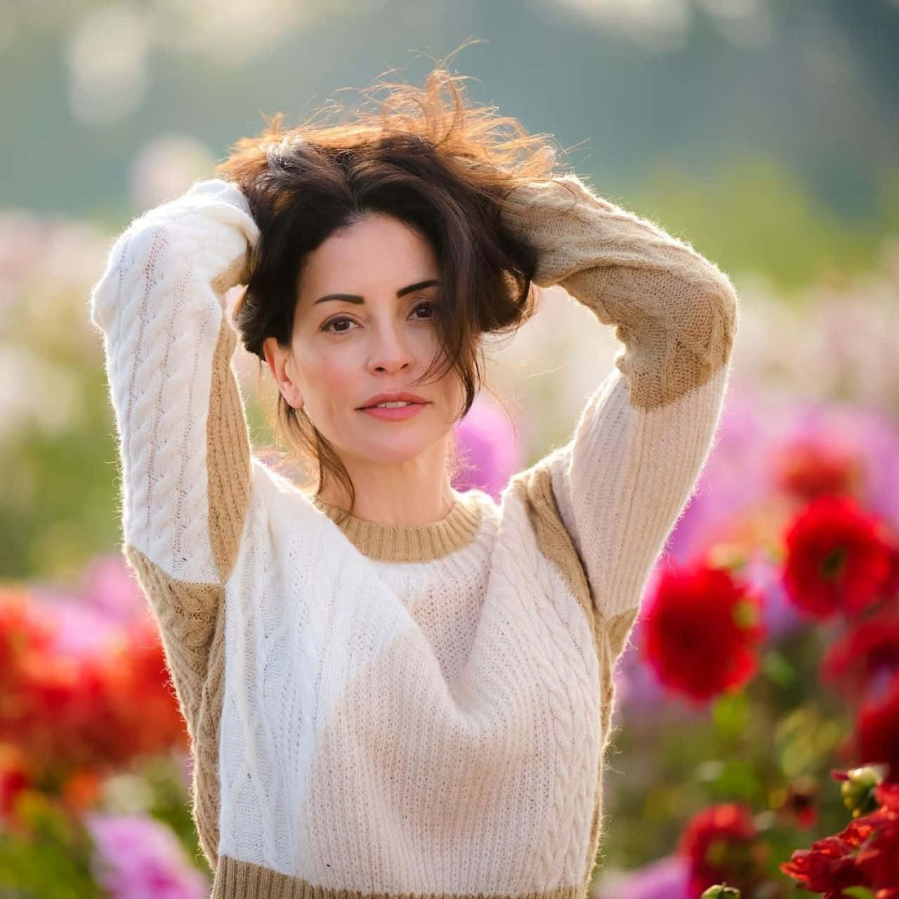 Emmanuelle Vaugier 桌布 壁紙 — 一位身穿奶油色麻花針織毛衣的女性，在一片鮮豔的紅色和粉色花田中，用手撥弄著她的深色頭髮。