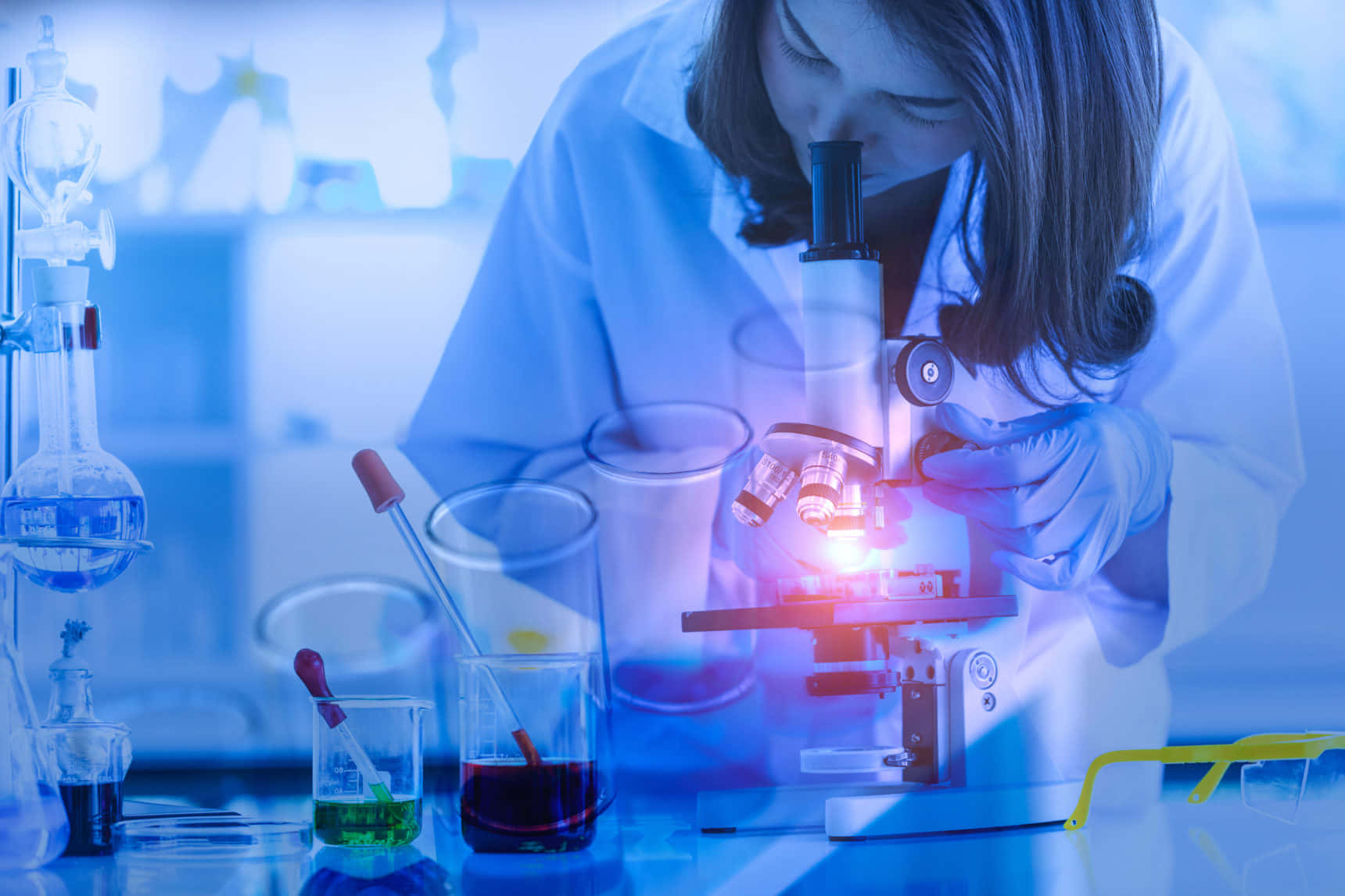 科學 桌布 壁紙 — 一位身穿實驗室外套的科學家專注地透過顯微鏡觀察，發光的紅光照亮了她專注的表情。