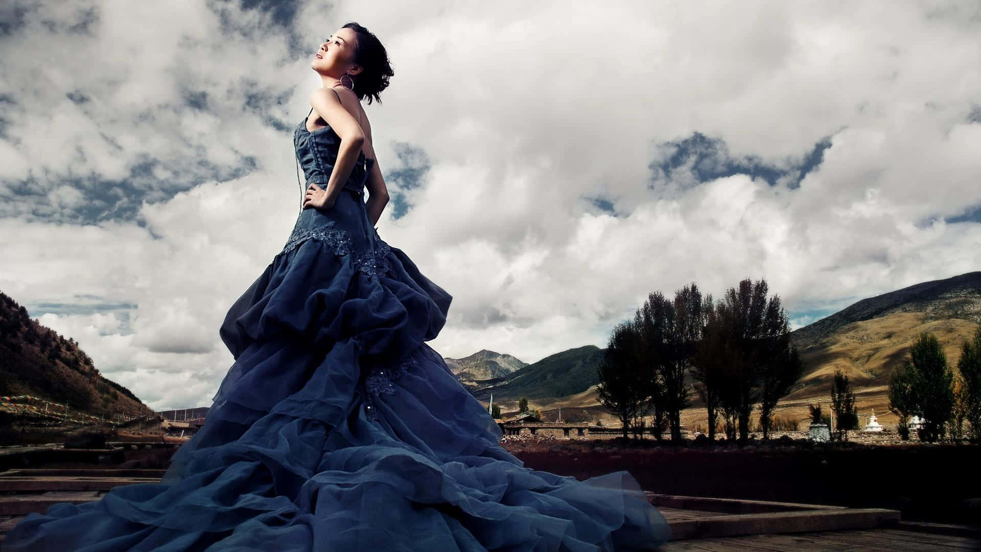 Navy Blue Dress 桌布 壁紙 — 一位身穿寬鬆深藍色長裙的女性，在戲劇性的多雲天空下，背景是連綿起伏的山丘和樹木，優雅地站立著。