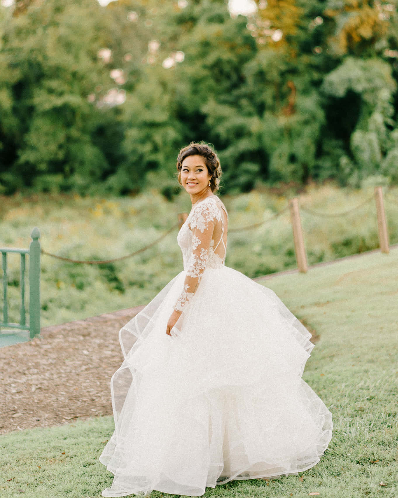 Bride 桌布 壁紙 — 一位身穿寬鬆白色蕾絲禮服的新娘，在陽光明媚的草地上，被茂密的綠樹環繞，回頭微笑。
