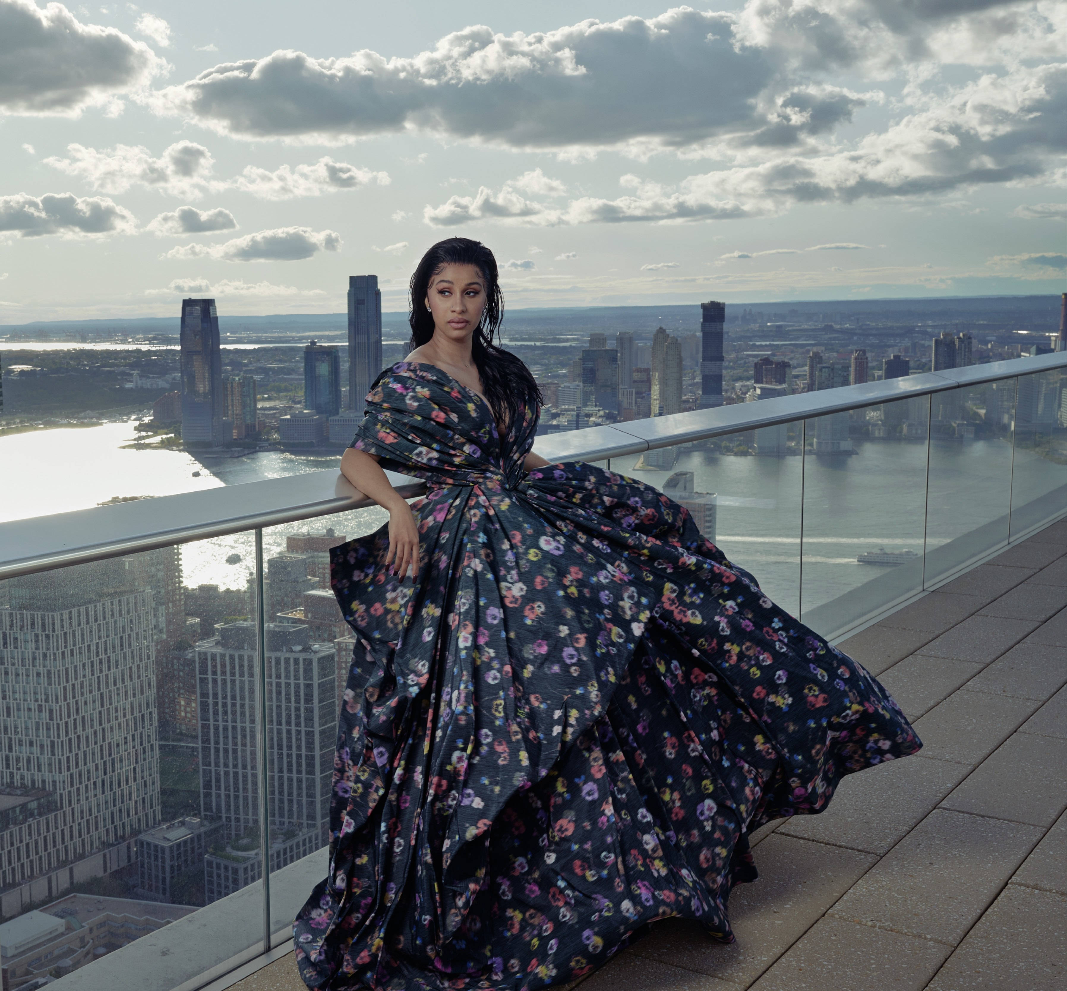 cardi 桌布 壁紙 — 一位身穿寬鬆花卉長裙的女性，站在玻璃欄杆的屋頂上，俯瞰著雲層下廣闊的城市景觀。
