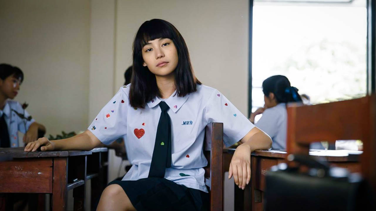 Nanno 桌布 壁紙 — 一位身穿帶有心形補丁校服的年輕女子，在陽光明媚的教室裡，若有所思地坐在木桌前。