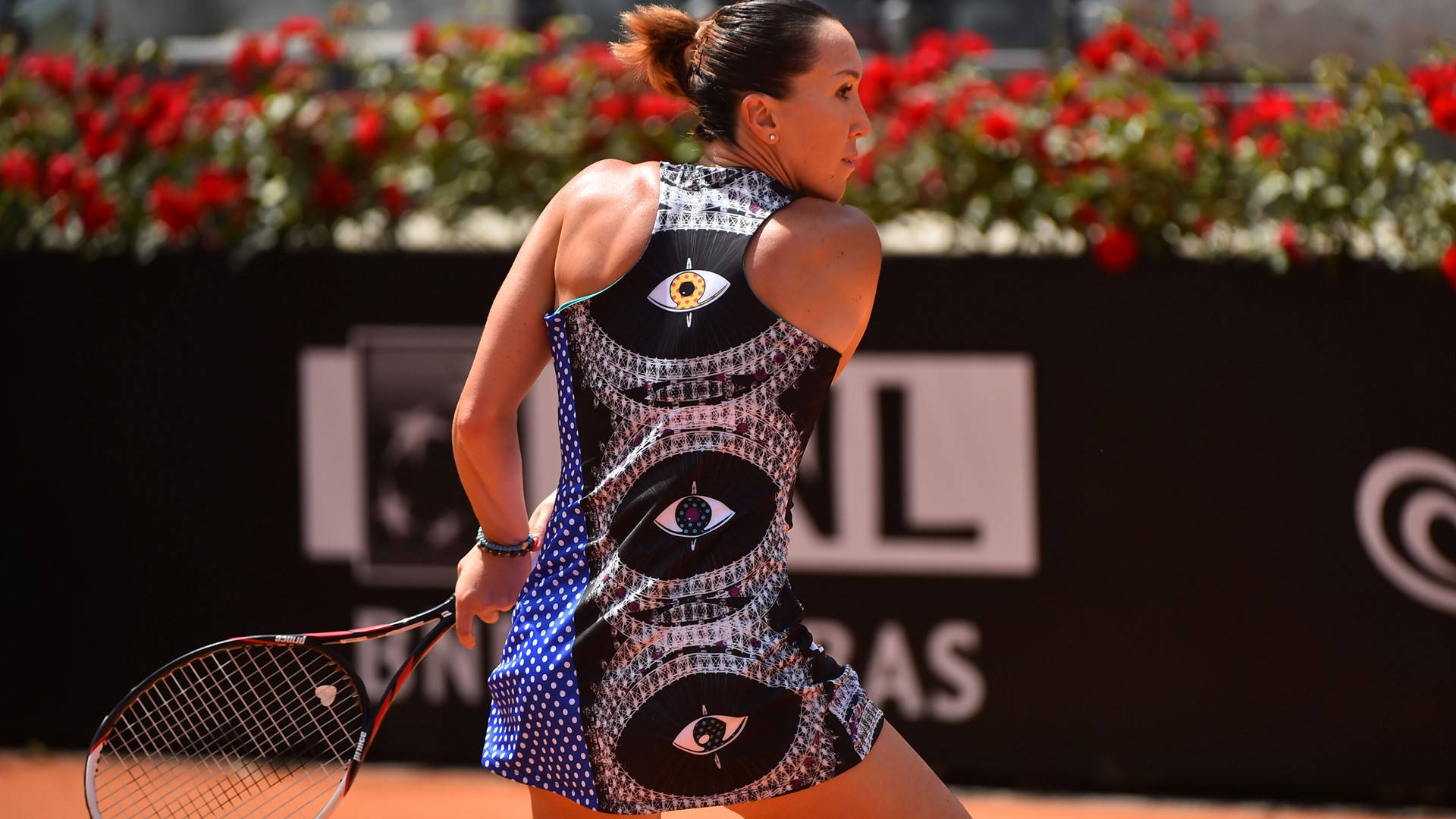 Tennis 桌布 壁紙 — 一位身穿帶有眼睛圖案連衣裙的女網球運動員，準備在紅花環繞的紅土場上擊球。